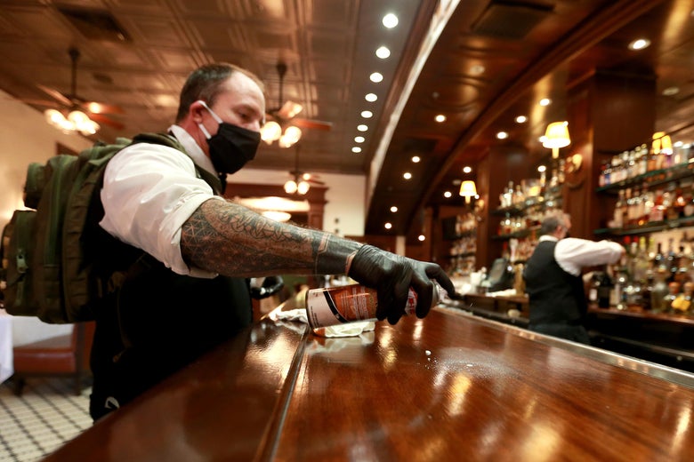 A bartender wearing a black mask and a backpack cleans the bar with disinfectant spray, while a bartender in the back mixes a drink.