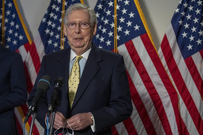 Mitch McConnell stands behind a microphone and in front of American flags while holding a face mask.