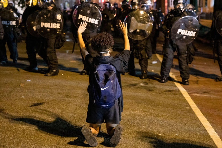 A young person with a backpack on kneels with their hands up in front of a line of police officers in riot gear.