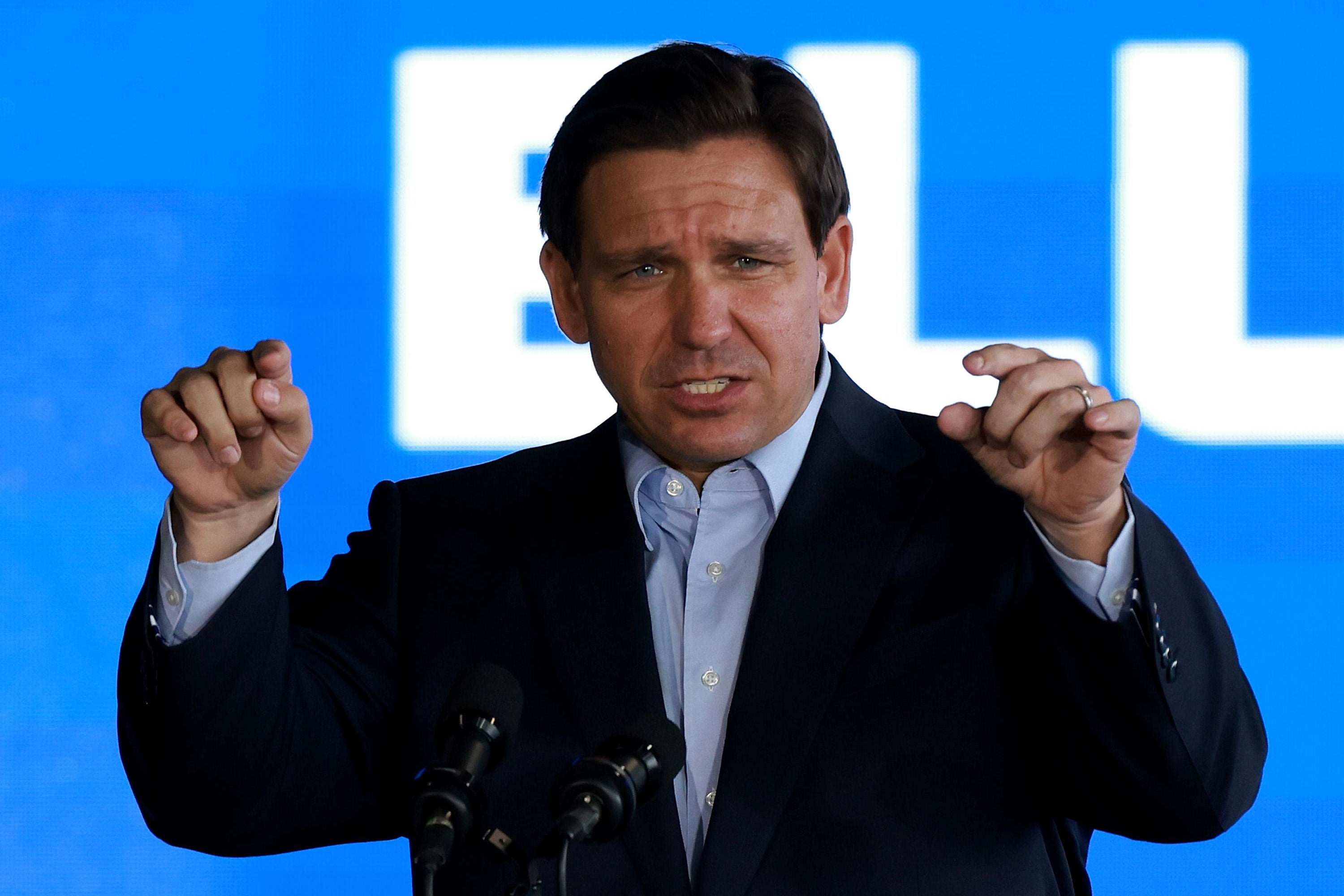 DeSantis gestures both of his hands in some sort of a half point, standing before a mic.