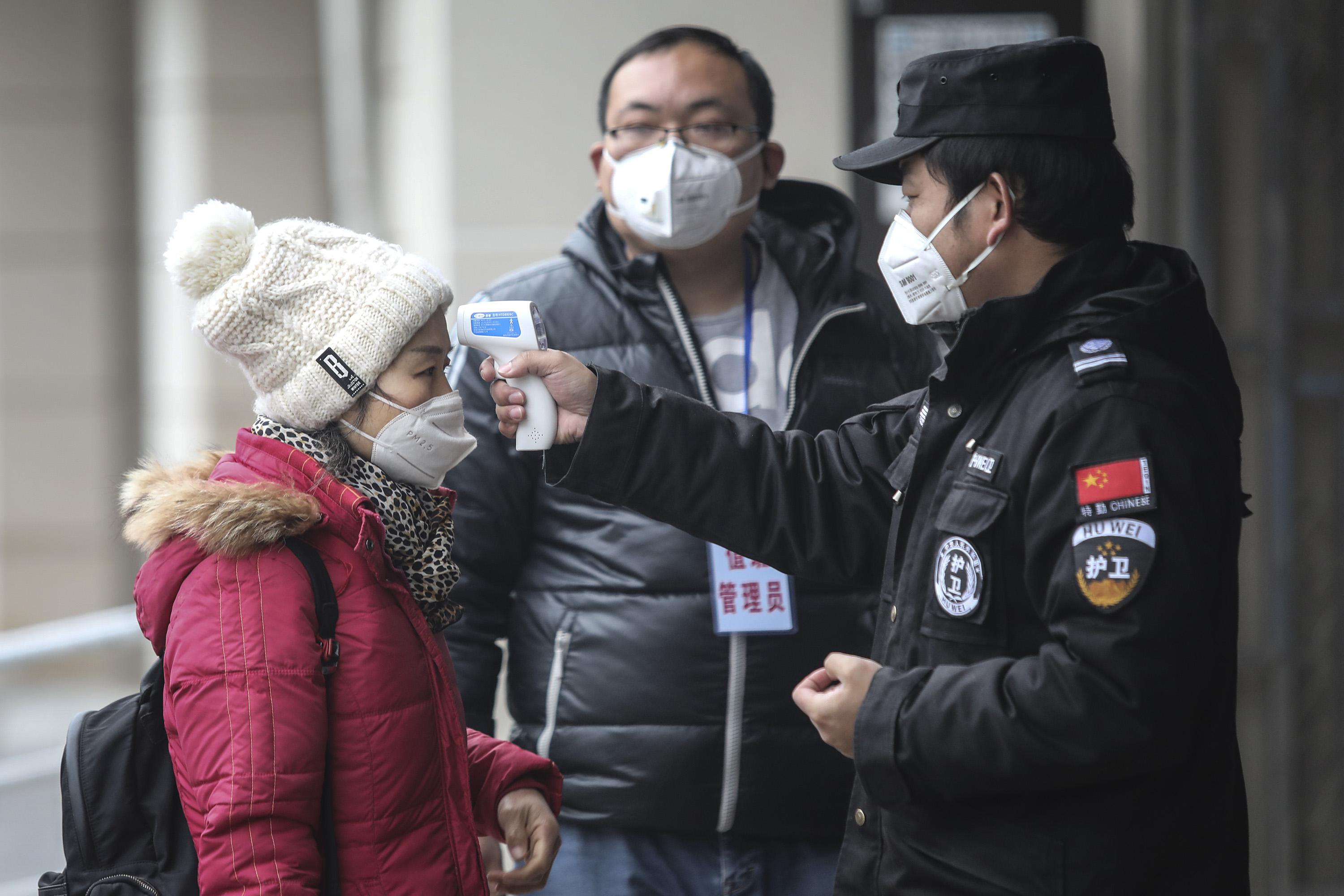 A security personnel on the right checks the temperature of a woman on the left. All 3 people in the photo are wearing flu masks. 