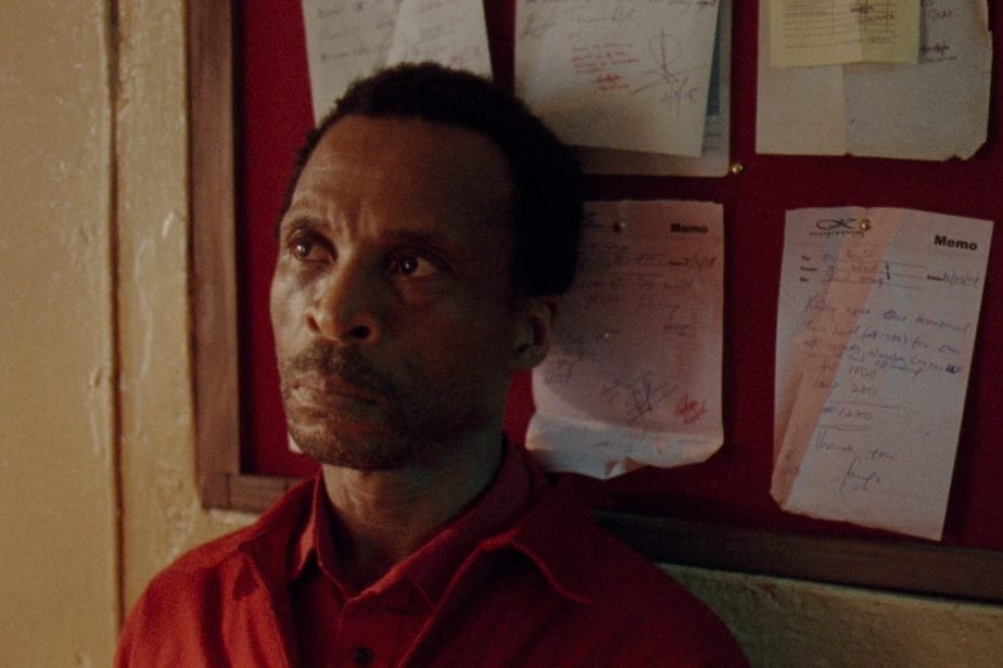 A Black man stands in front of a bulletin board in a red polo while looking up and away from the camera with a straight face.