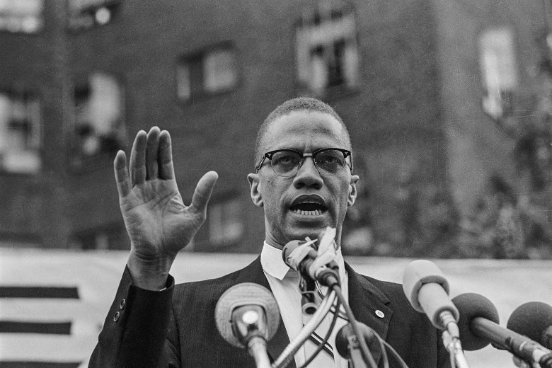 Malcolm X raises his right hand as he stands outside a building speaking into a cluster of microphones