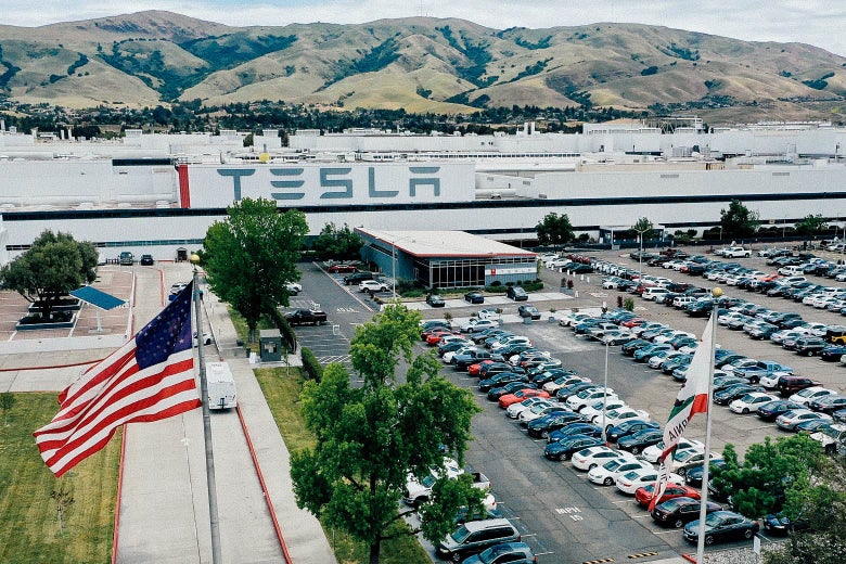 A Tesla factory with many cars parked in the parking lot. There are mountains behind it.