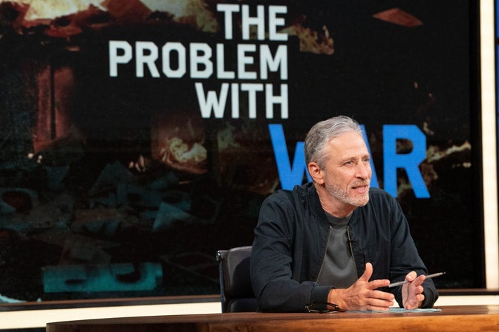 Jon Stewart sits at a desk and gesticulates in front of a display reading, "The Problem With War."