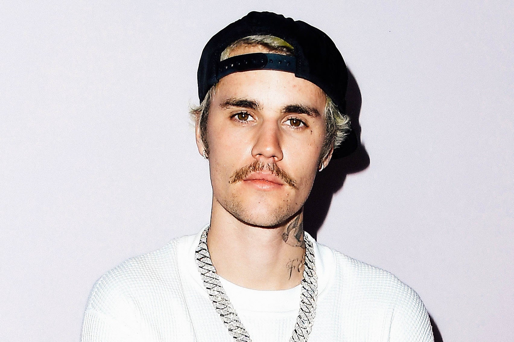 Justin Bieber in a white shirt, black hat, and silver chain, sporting a mustache.
