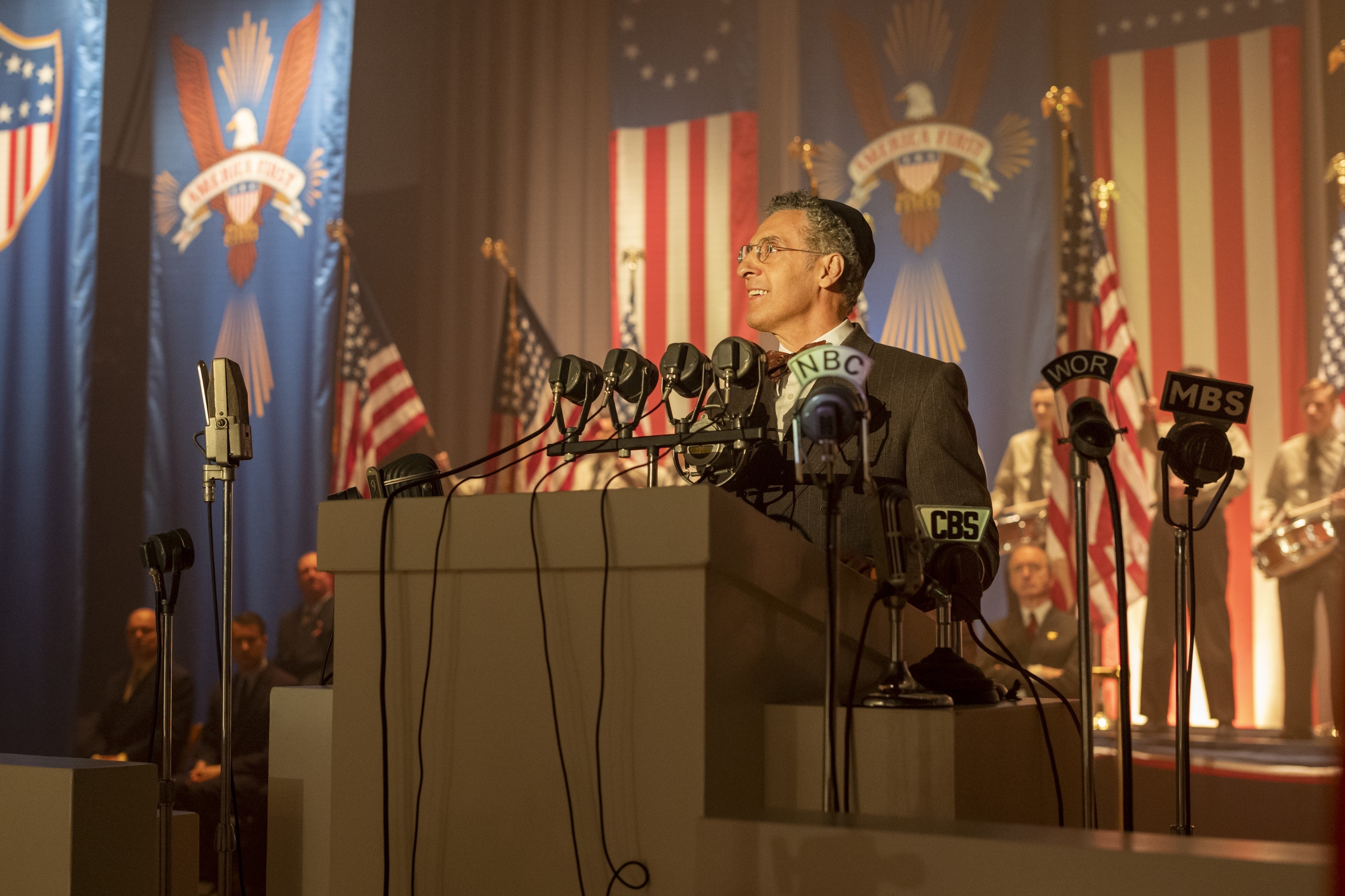 John Turturro stands at a podium, surrounded by microphones and American flags.