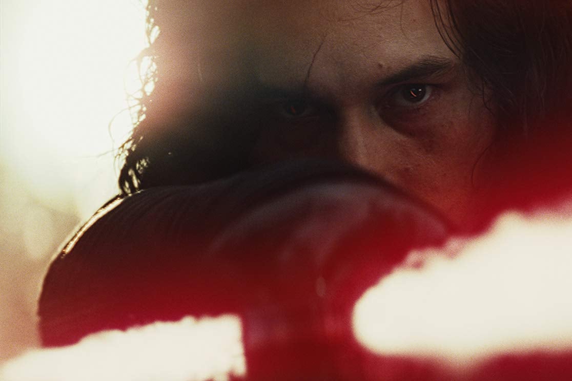 A close up of Adam Driver as Kylo Ren, peering over the red light of a lightsaber.