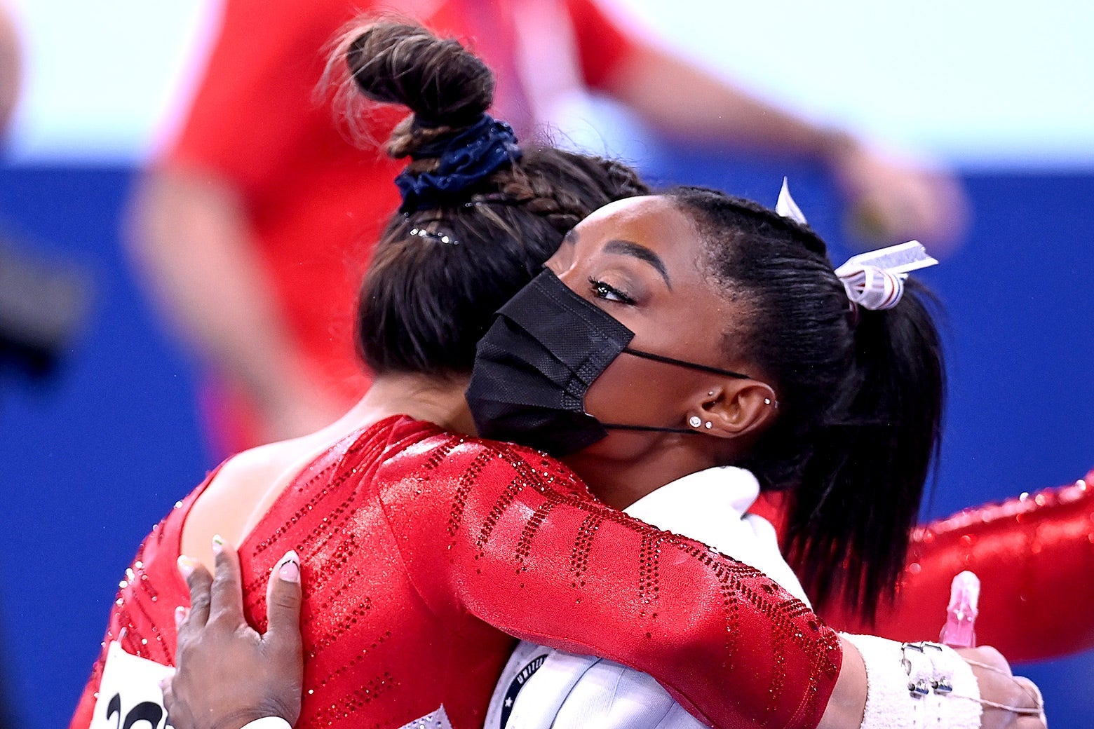 Biles wearing her warmup suit and a black mask hugs Sunisa Lee wearing a competition leotard