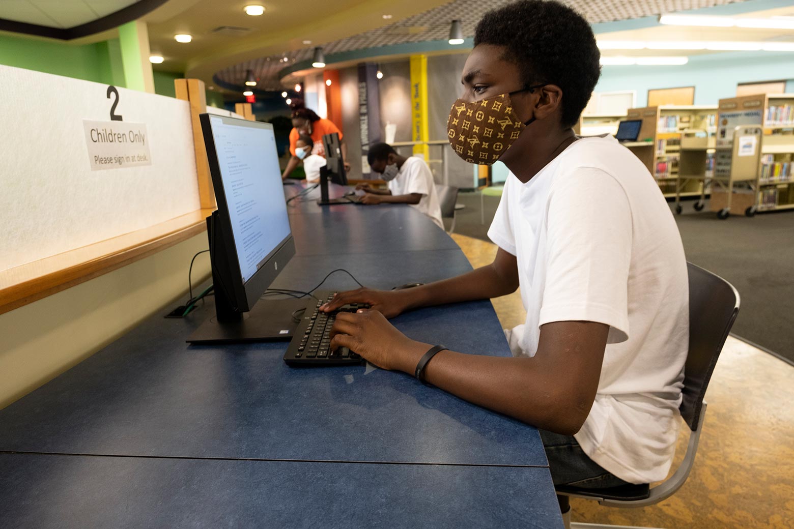 Teenage boy wearing a face mask working at a desktop computer in a public library. In the background, one of his brothers works at a different computer, and his mother stands beside the third brother, all three wearing masks.