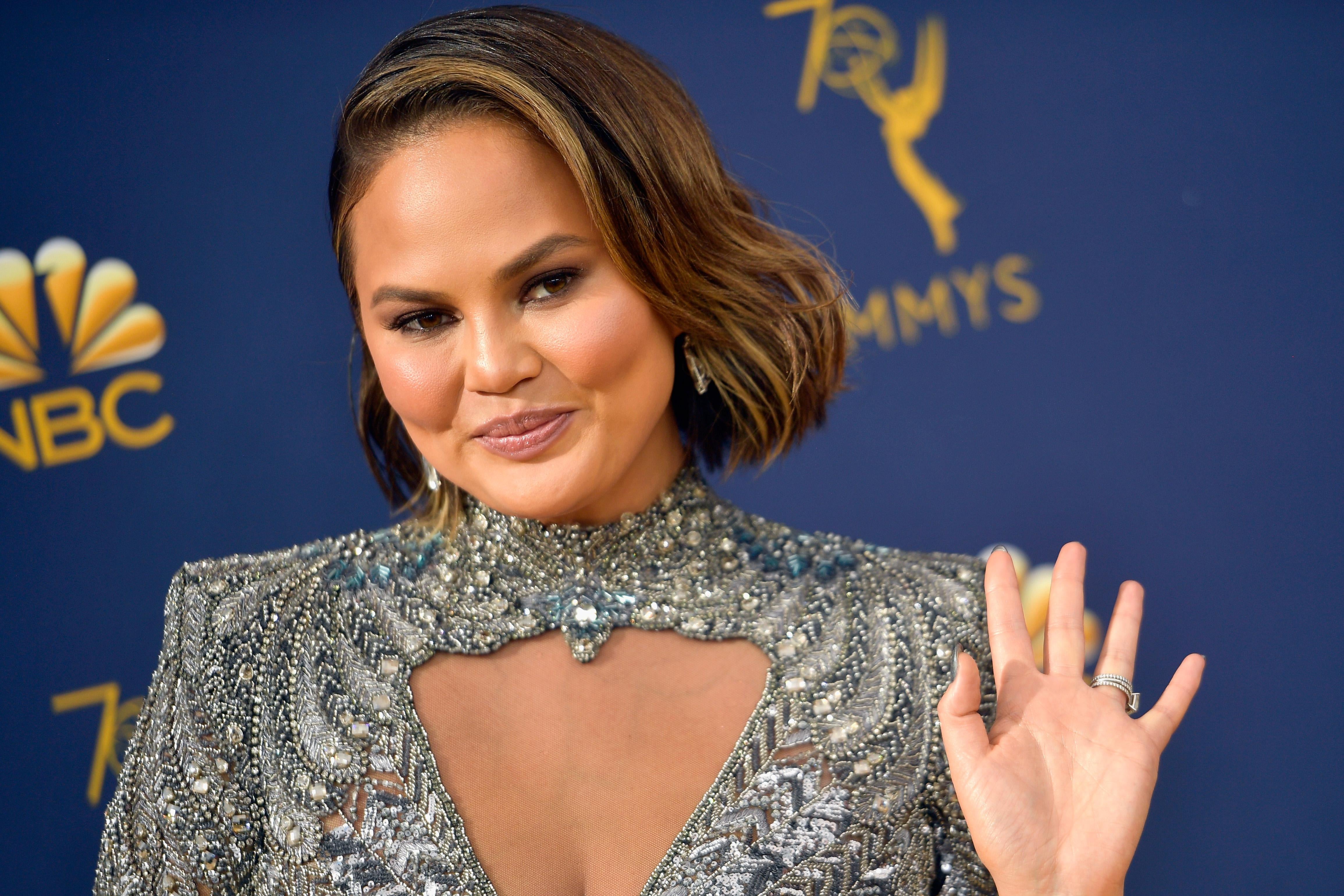 Chrissy Teigen waves at the camera, wearing a silver high-neck gown. 