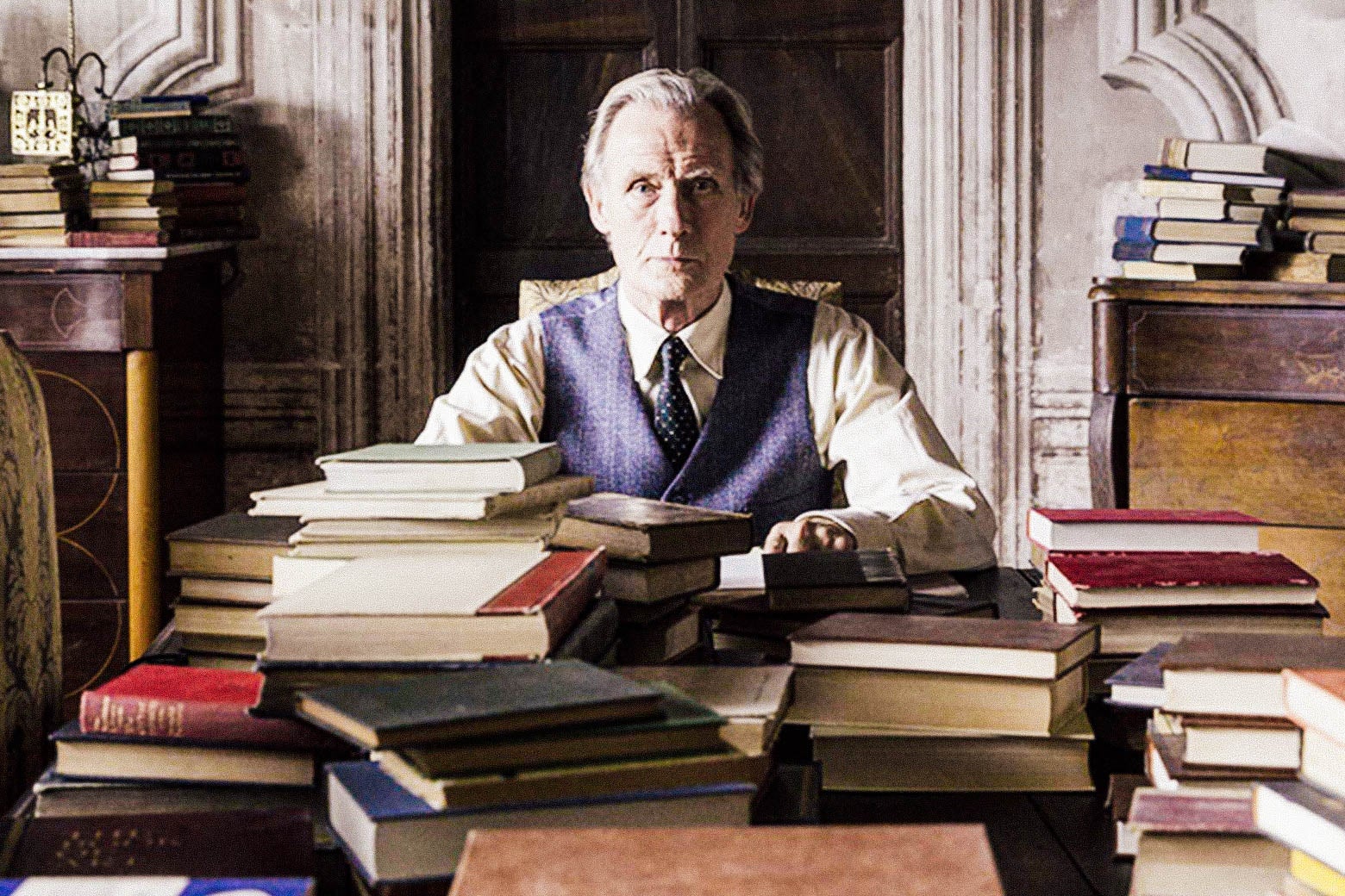 In a still from The Bookshop, Mr. Brundish (Bill Nighy) sits at a desk covered in and surrounded by books.