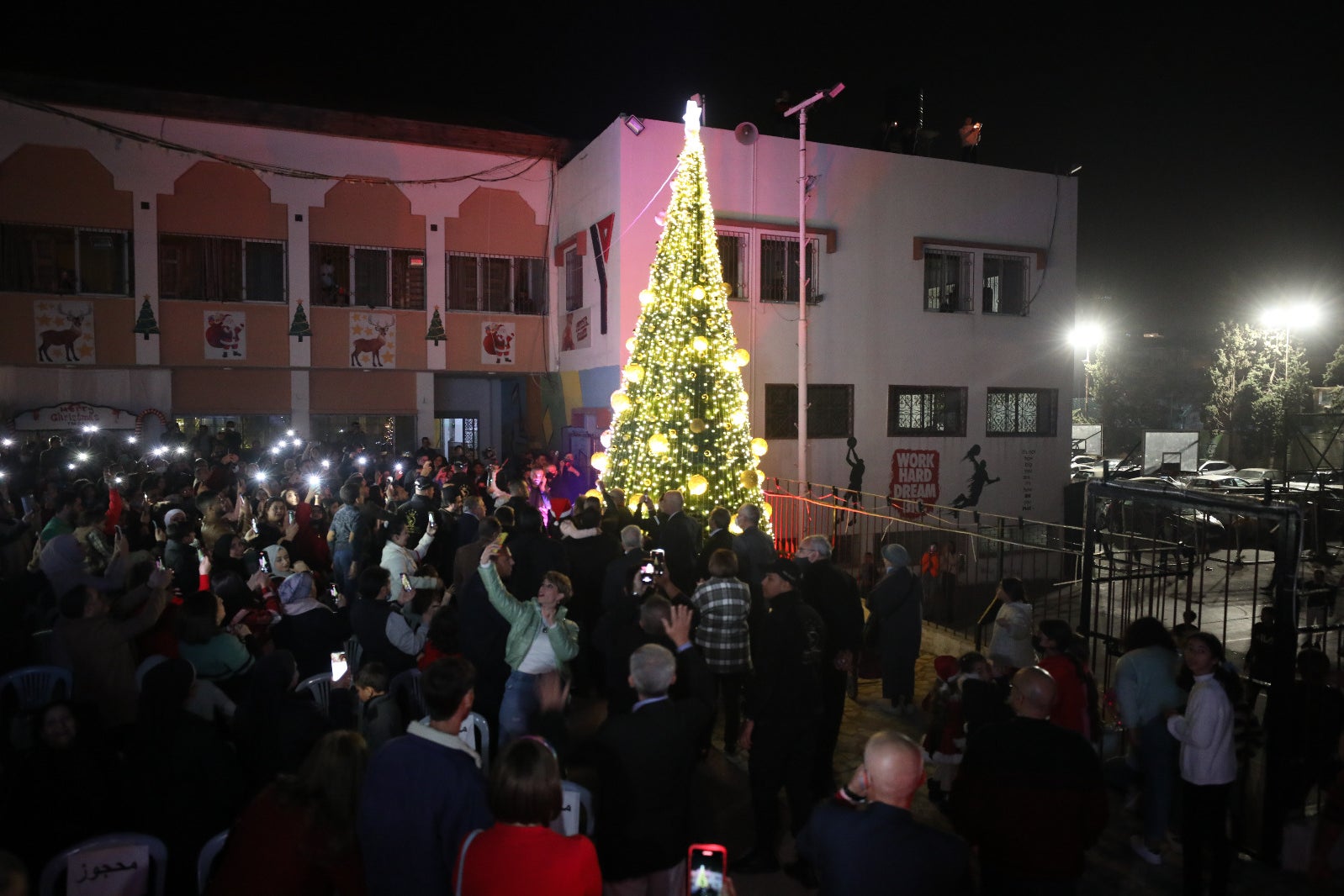 A crowd takes their selfies, many with flash on, in front of the big lit up Christmas tree at the YMCA.