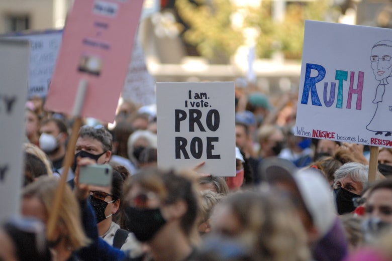 Protesters march outside holding signs including one that says "I am. I vote. Pro Roe" and one with a drawing of Ruth Bader Ginsburg