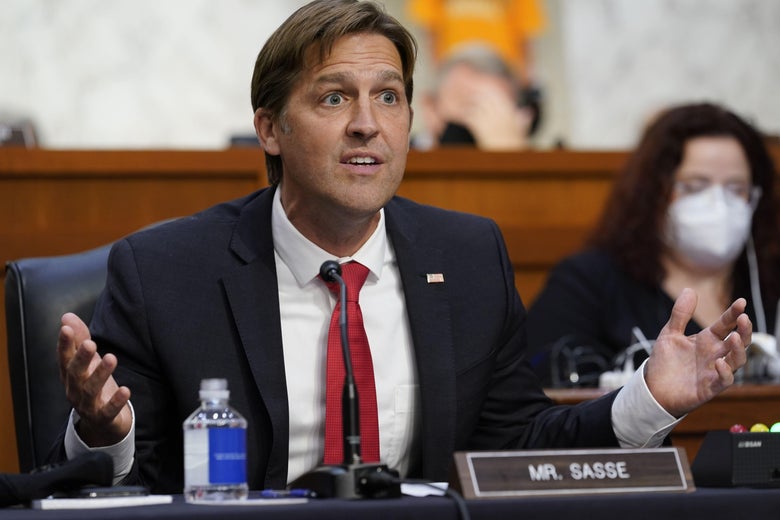 Ben Sasse, in a dark suit and red necktie, is seen from his Senate desktop up as he sits and gesticulates with both hands, exposing extra shirt sleeve below his suit cuffs. Over his left shoulder and out of focus is a woman wearing a white protective mask.