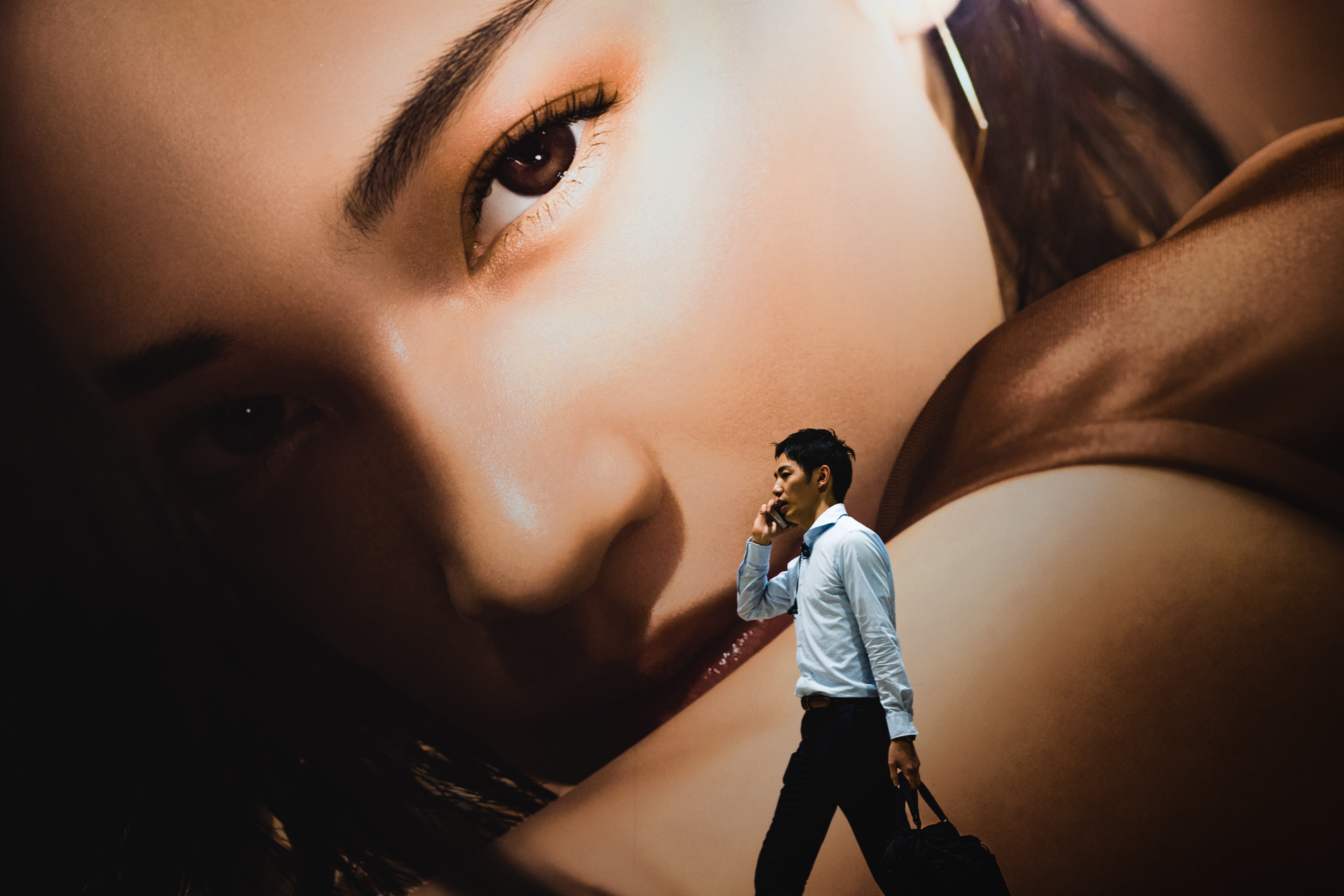 A man on a cellphone walks in front of an enormous image of a woman's face and arm.