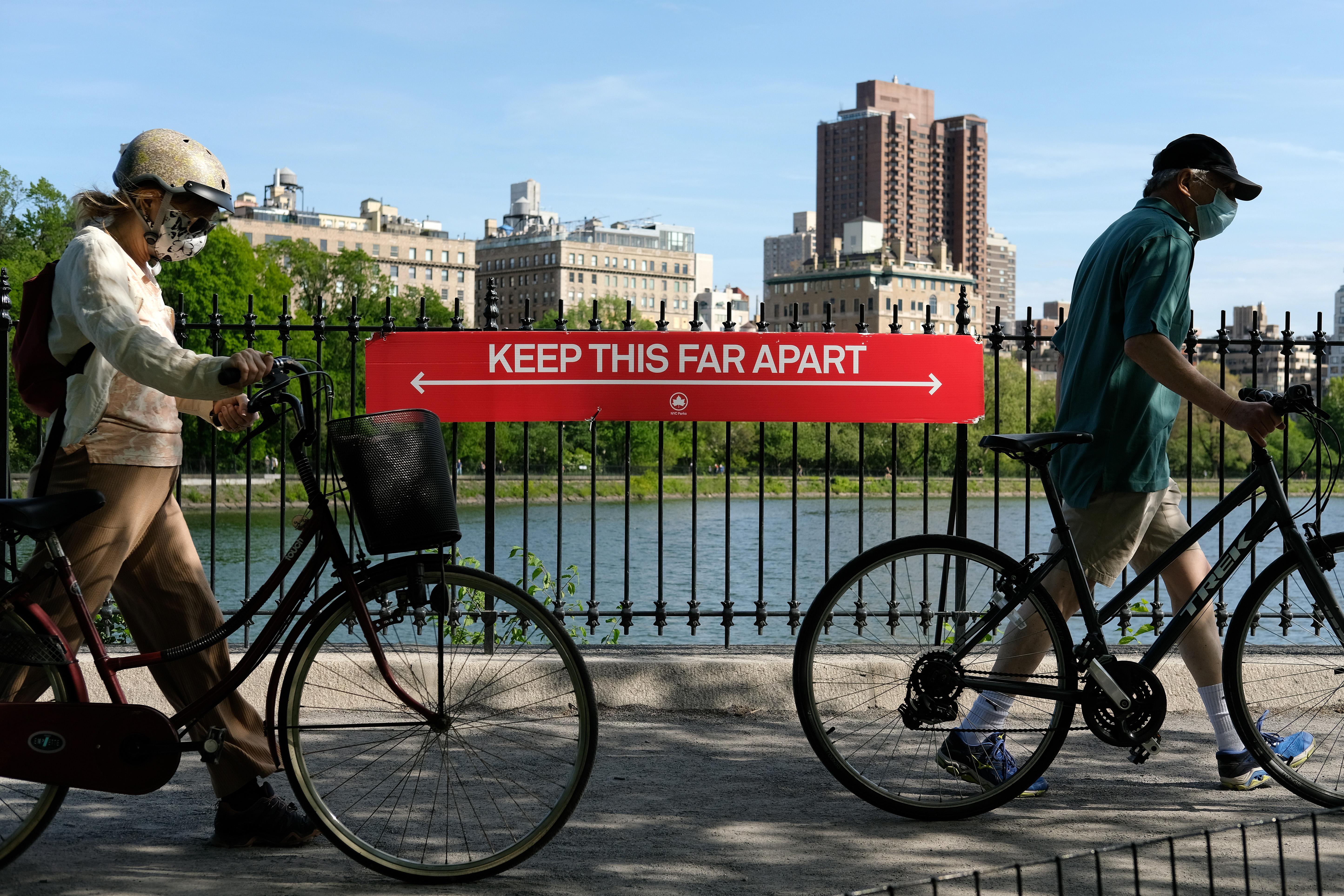 People wearing protective masks walk their bicycles past a social distancing sign reading "KEEP THIS FAR APART" in Central Park.