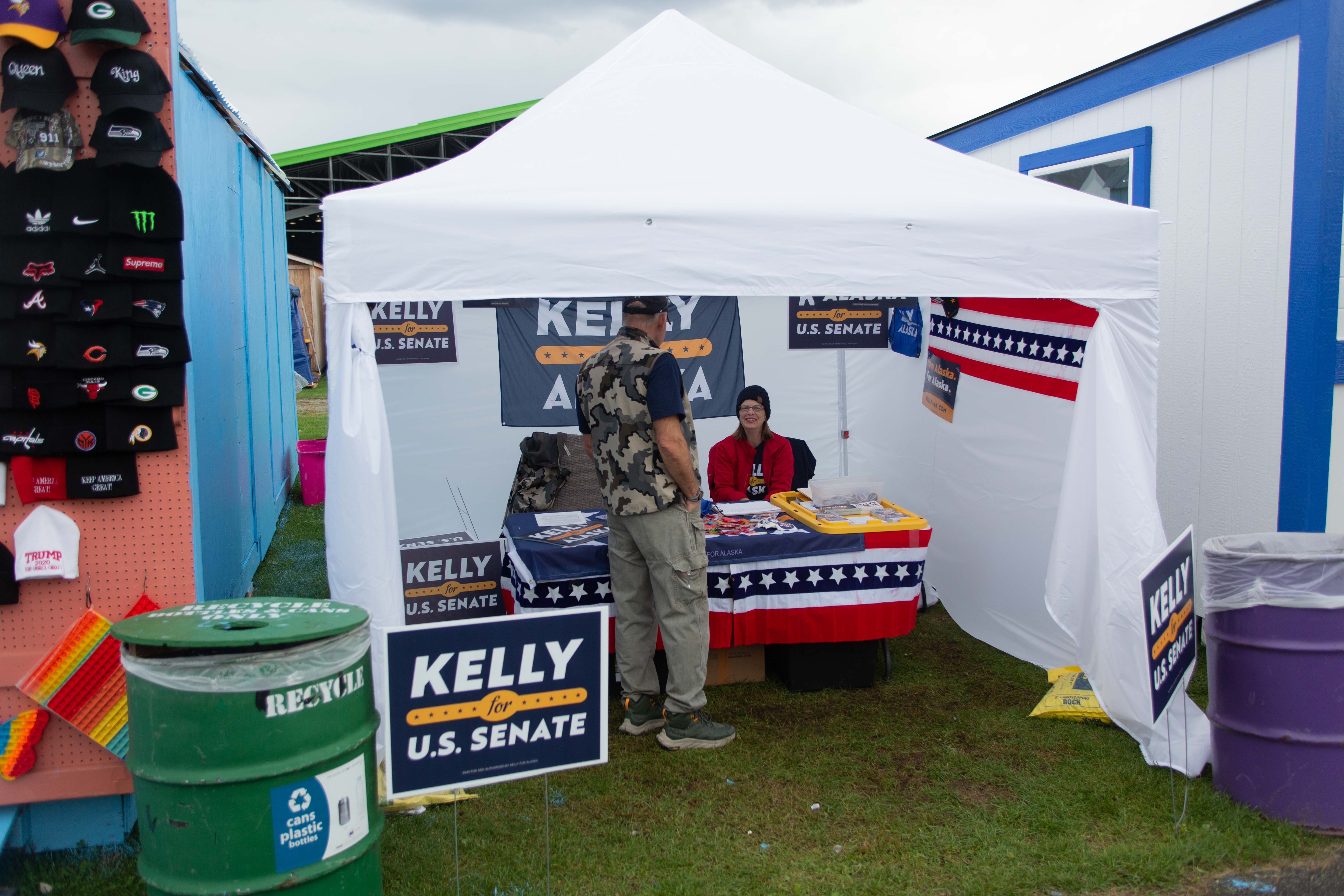 A tent with a man inside speaking with a woman, with signage around saying “Kelly for Senate”.