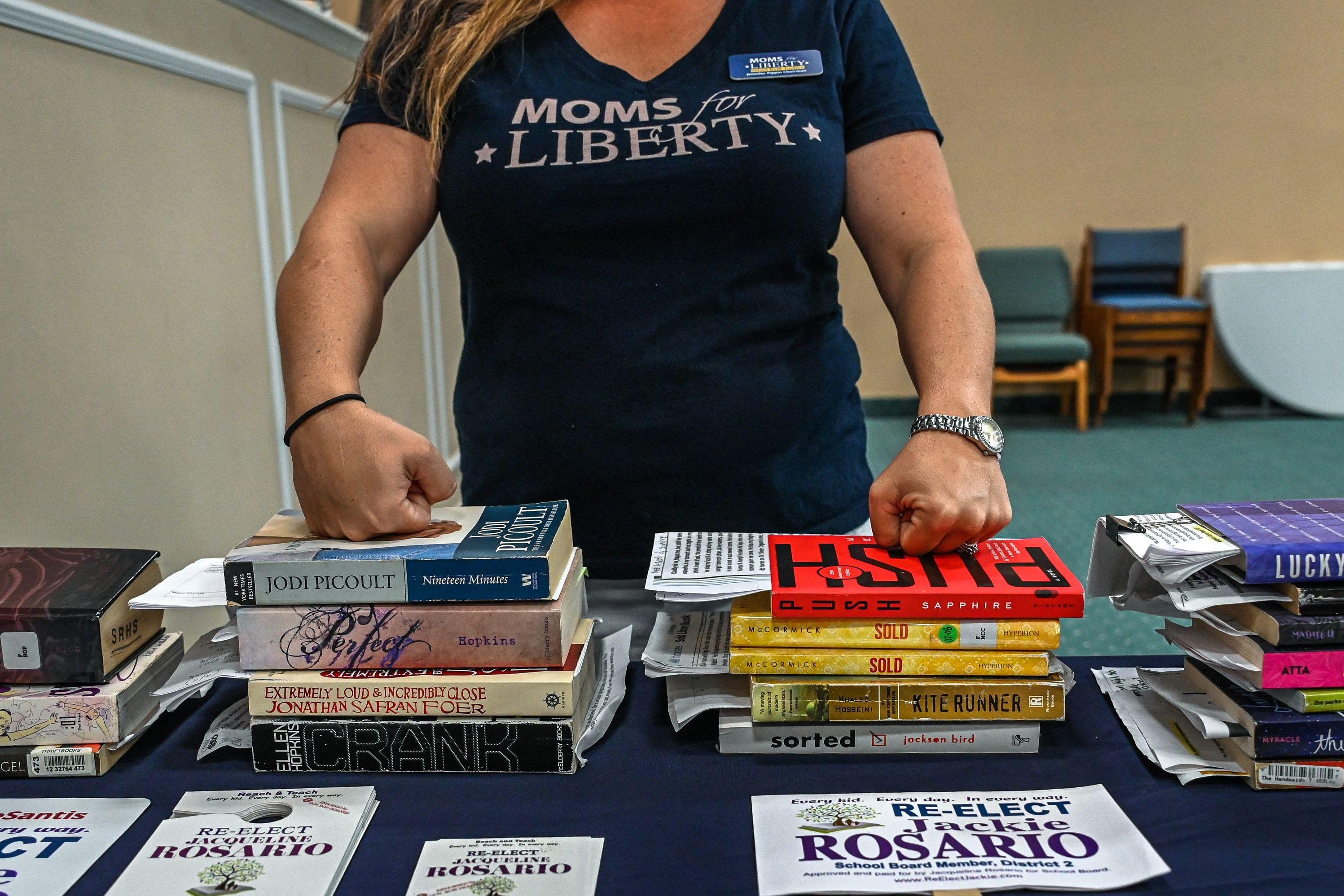 Pippin wearing a "Moms for Liberty" shirt and placing her hands over stacks of books.