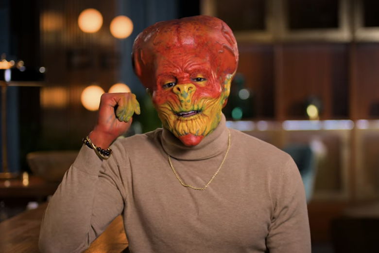 Man in beige sweater wearing a red and yellow alien head.
