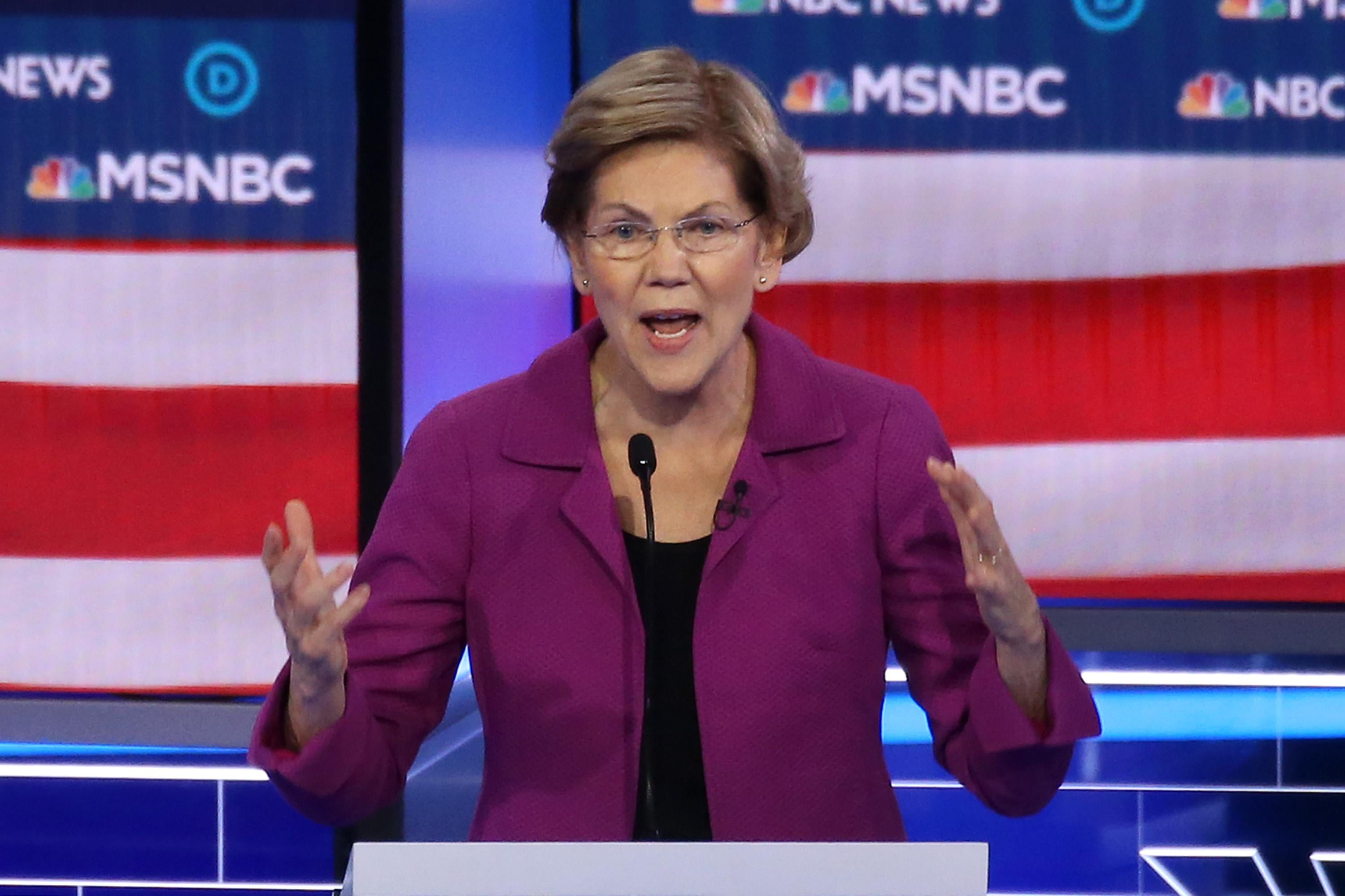 Elizabeth Warren raises her hands while speaking into a microphone.