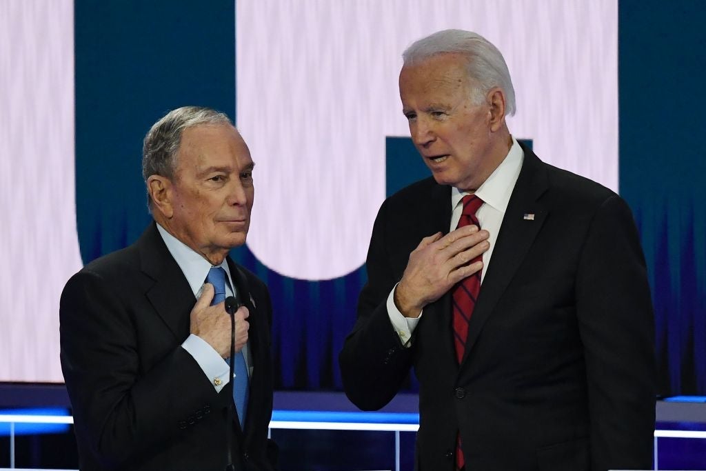 Bloomberg and Biden converse while holding their hands over the microphones that are affixed to their suits.