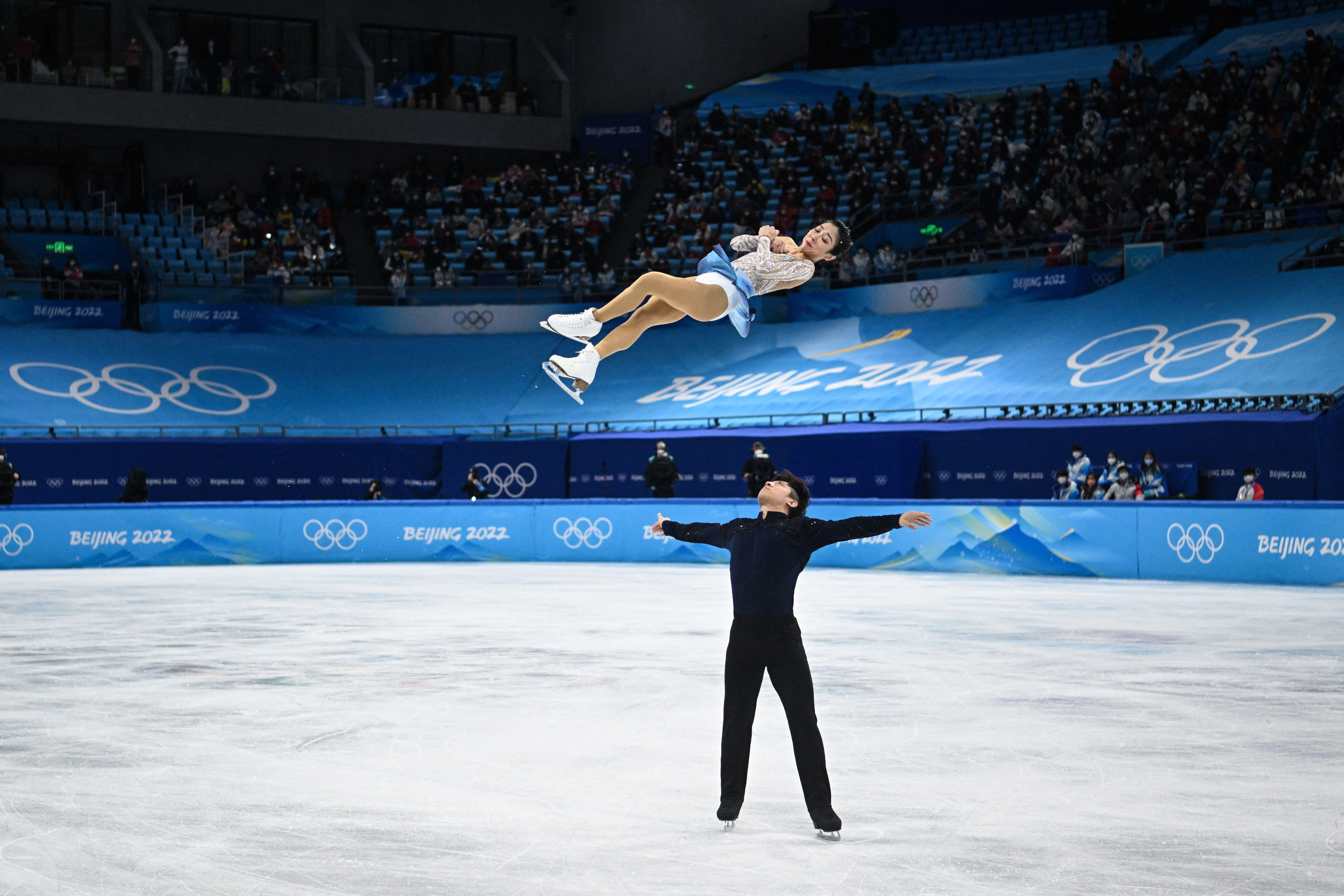 Sui spinning high and horizontally in midair, as Han watches her with his arms outstretched, after throwing her, prepared to catch, on the ice