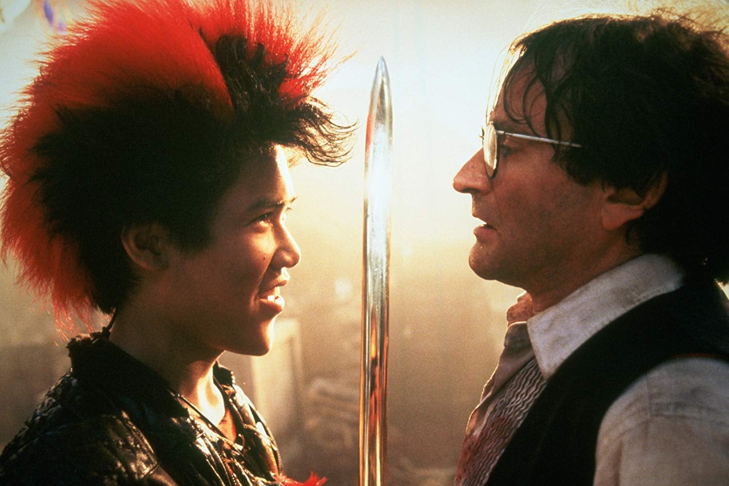 Dante Basco, with a red-tipped mohawk, holds a sword up to a bespectacled Robin Williams.
