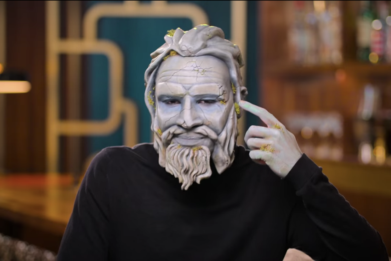 Man in black sweater pointing to his gray painted face.
