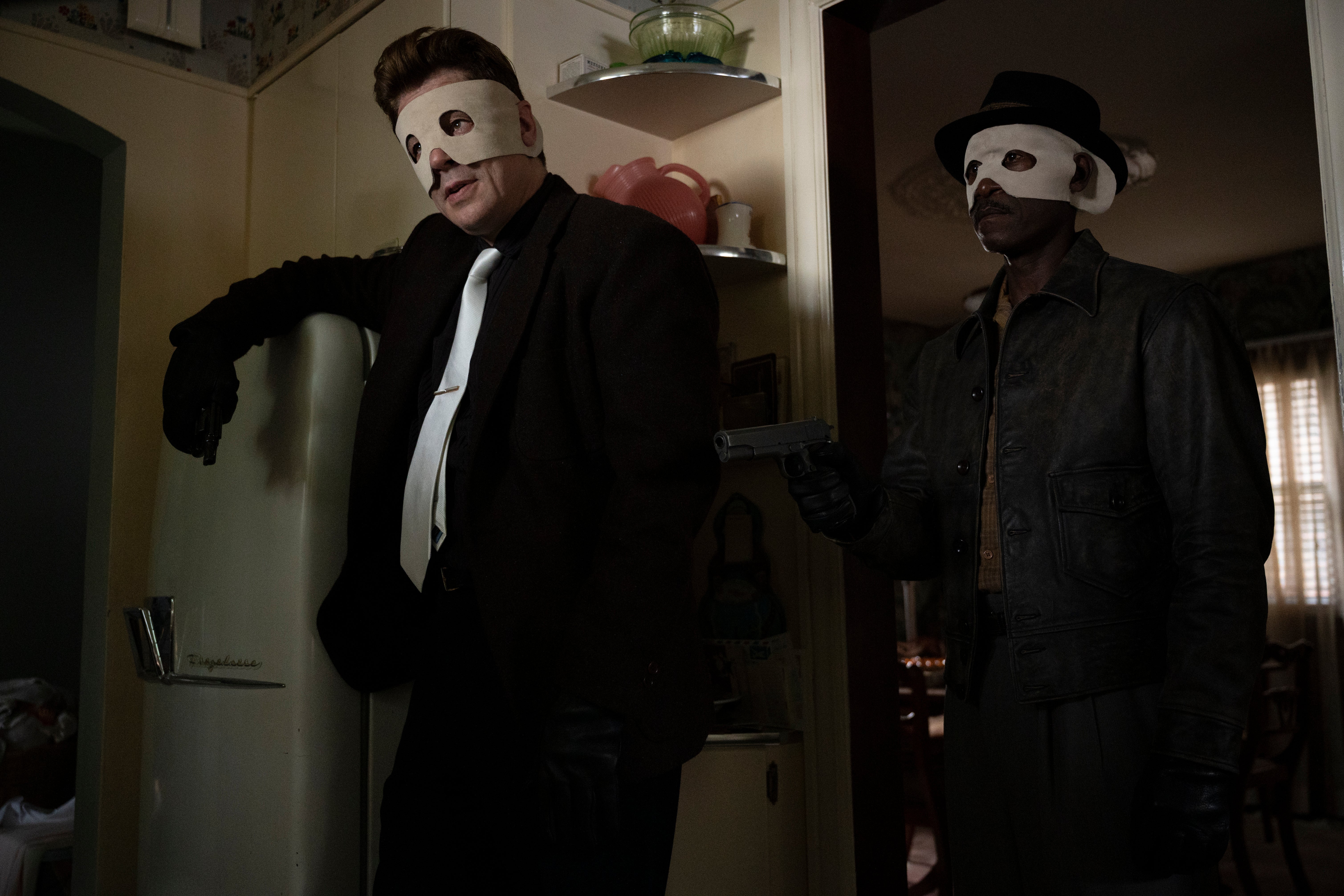 The two actors wear crude white masks and 1950s clothes in a 1950s kitchen