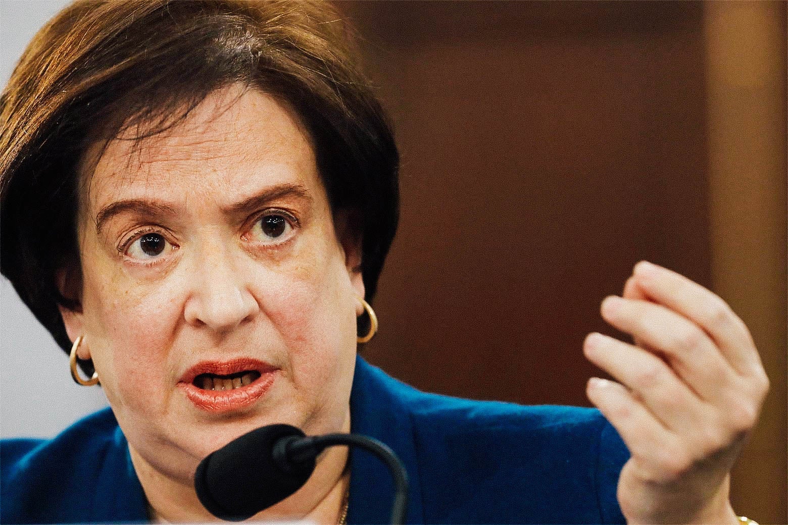 Elena Kagan speaks into a microphone and raises her hand.