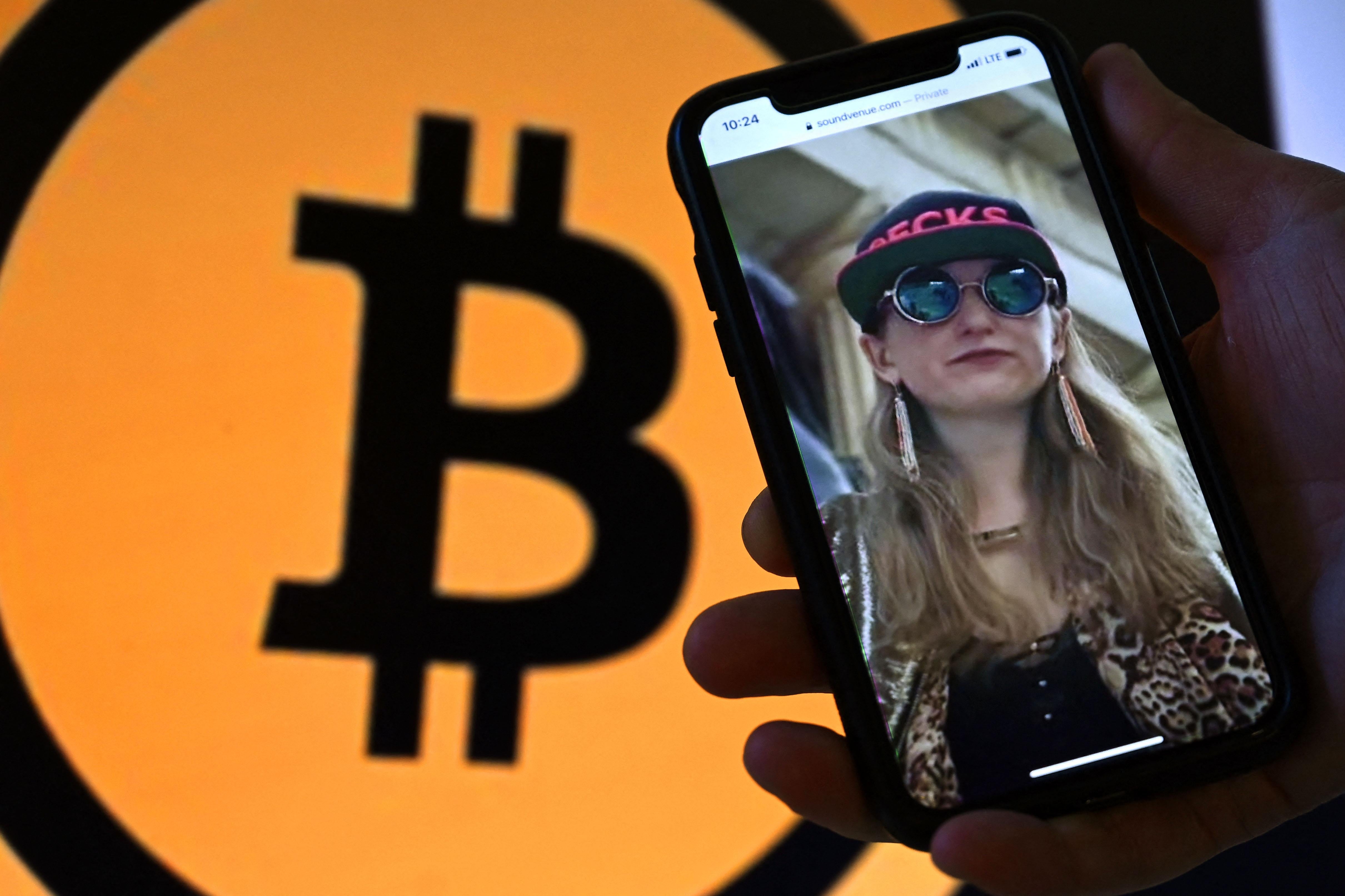 This illustration photo shows Heather Morgan, also known as “Razzlekhan,” on a phone in front of the Bitcoin logo displayed on a screen.