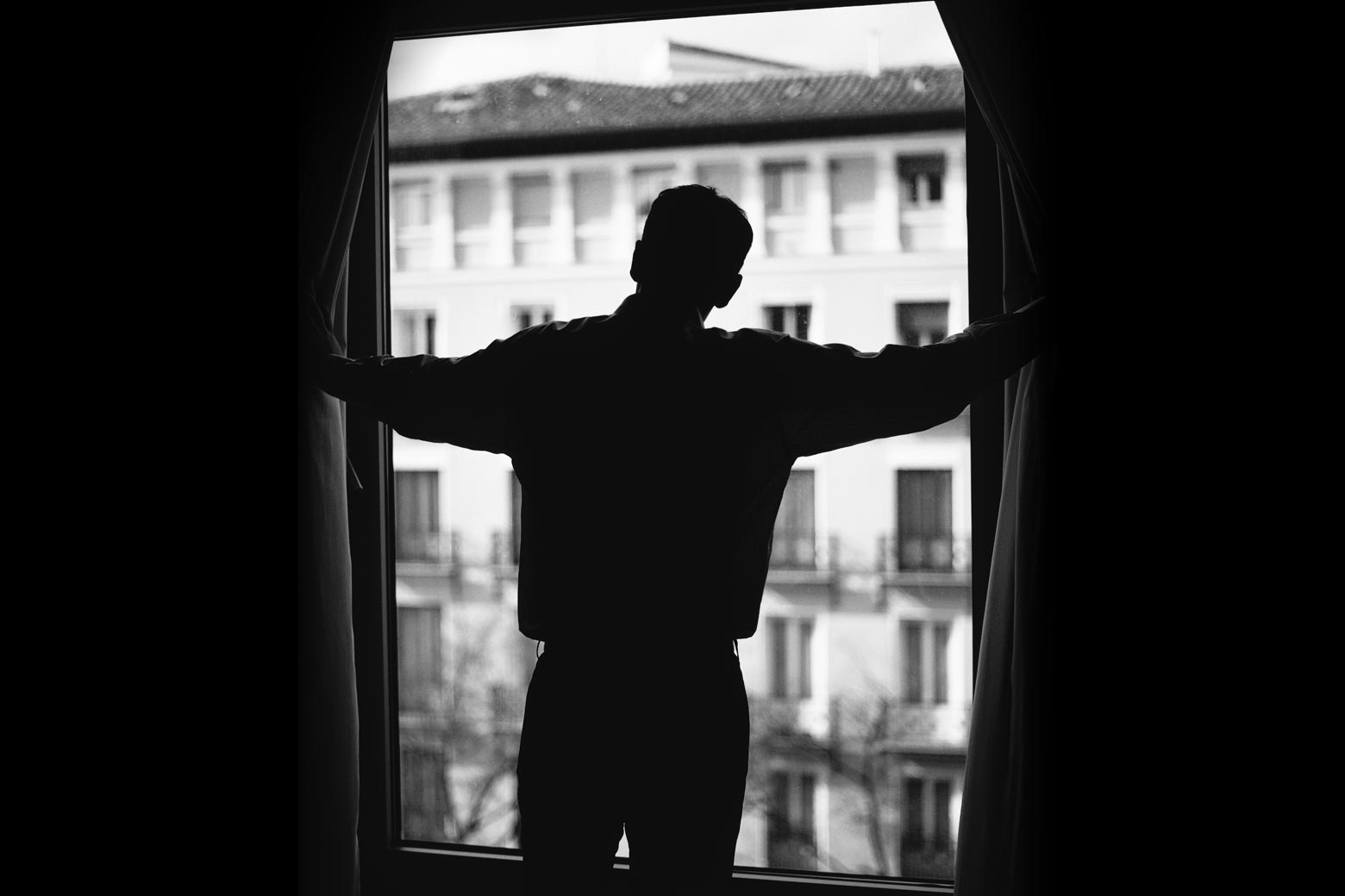 Man looking out of a window, pushing back the curtains.
