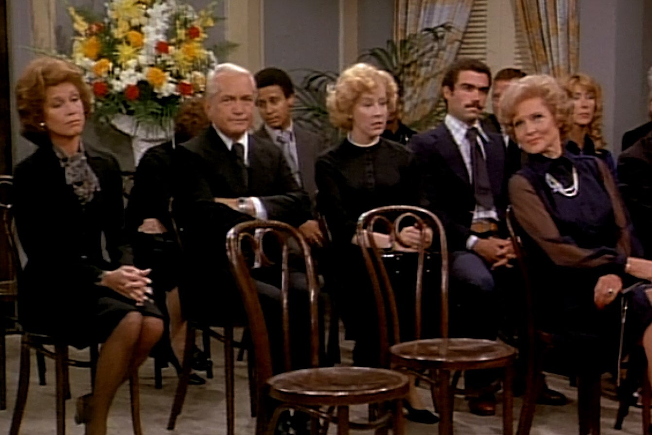 Attendees of a funeral, including Mary Tyler Moore and Betty White, sit in wooden chairs. In the front row, two chairs are empty.