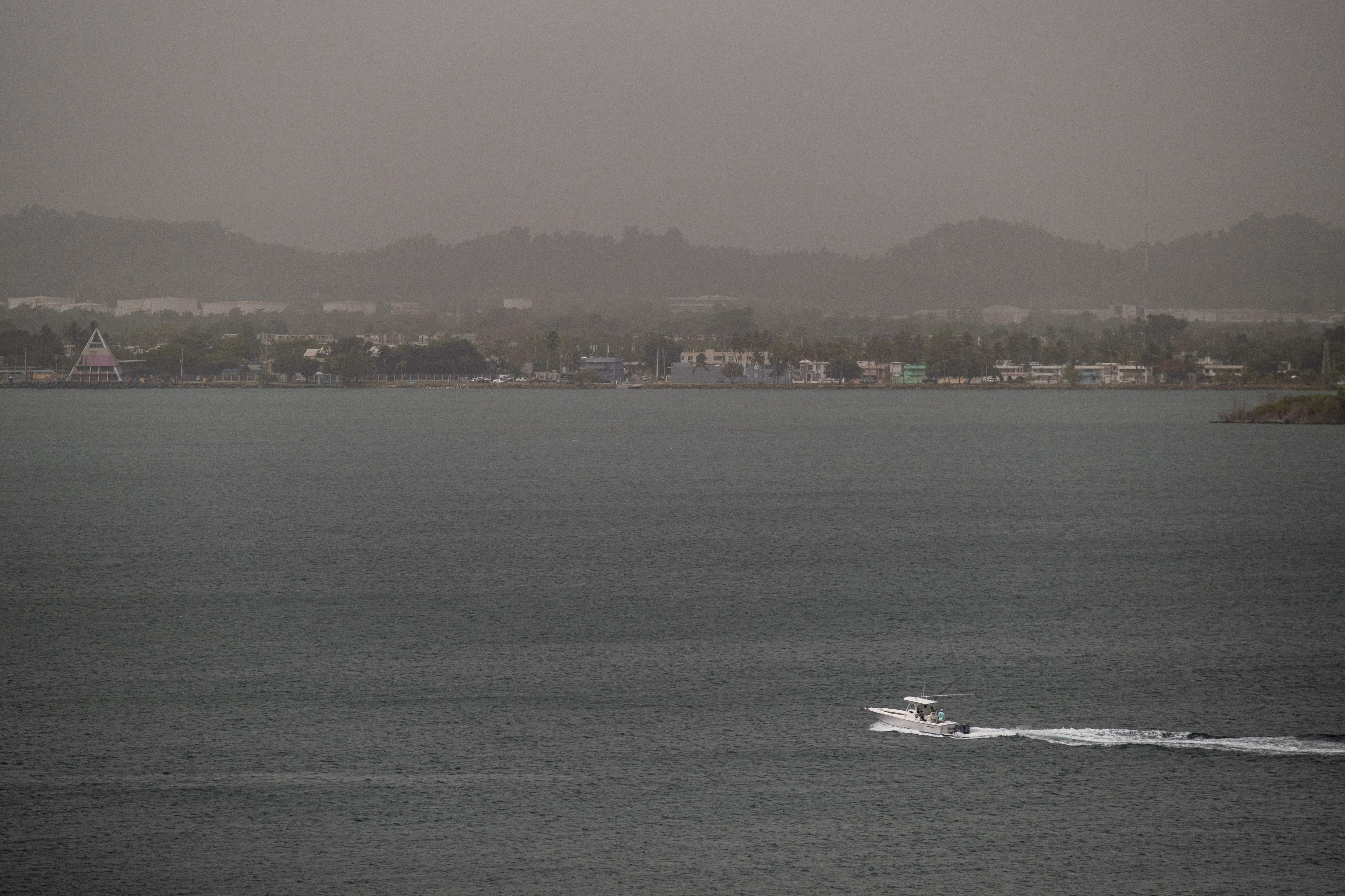 A boat speeds across a bay in front of a shoreline enveloped in a haze. The air is gray, and the mountains and buildings are barely visible.