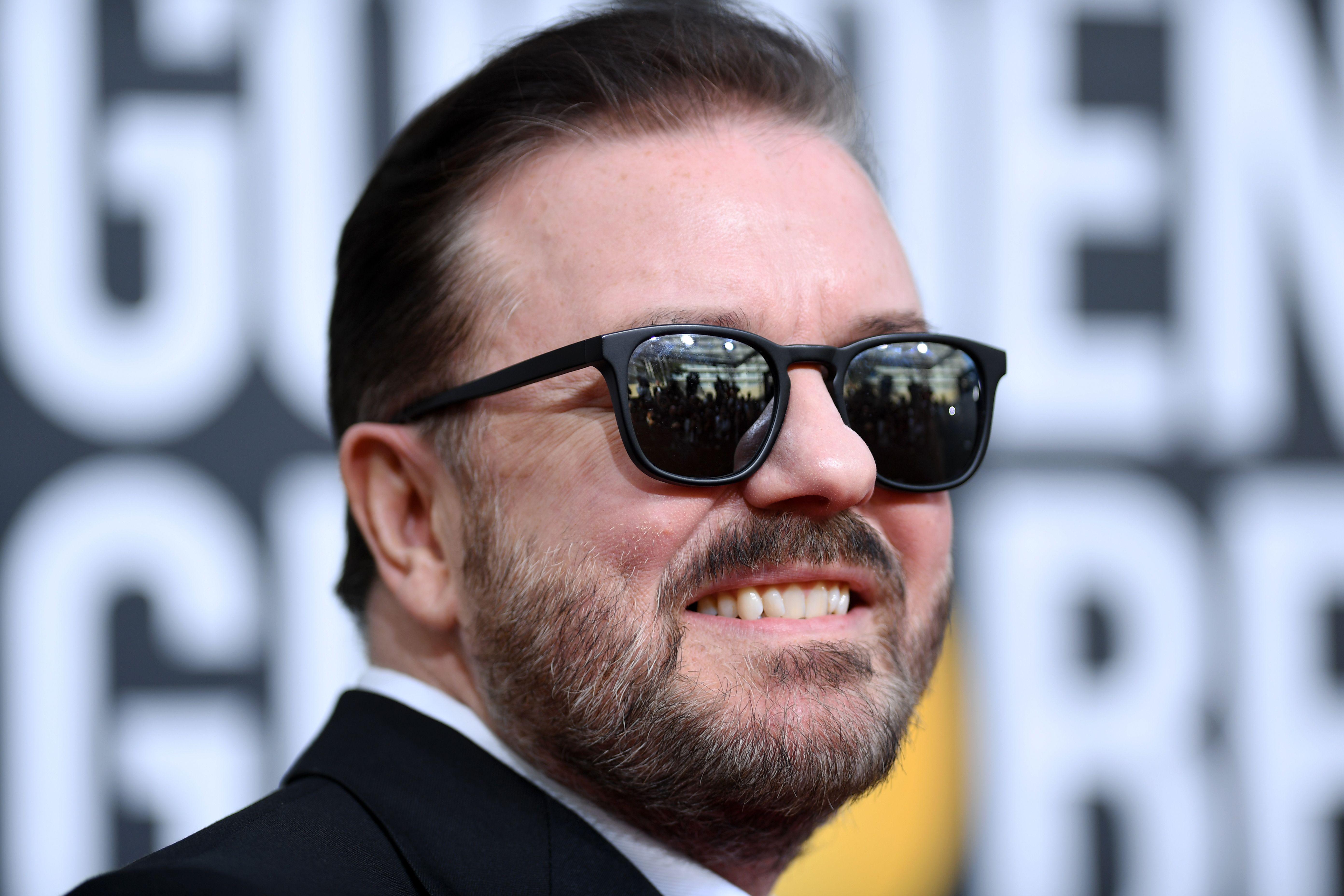 Ricky Gervais smiling in sunglasses with a shit-eating grin.