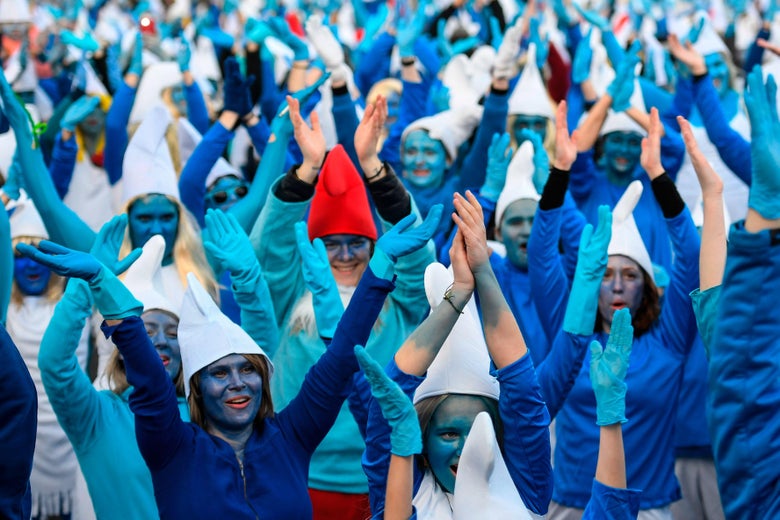 A crowd of people dress as Smurfs by wearing blue, painting their faces and arms blue, wearing blue gloves, and donning red and white caps.
