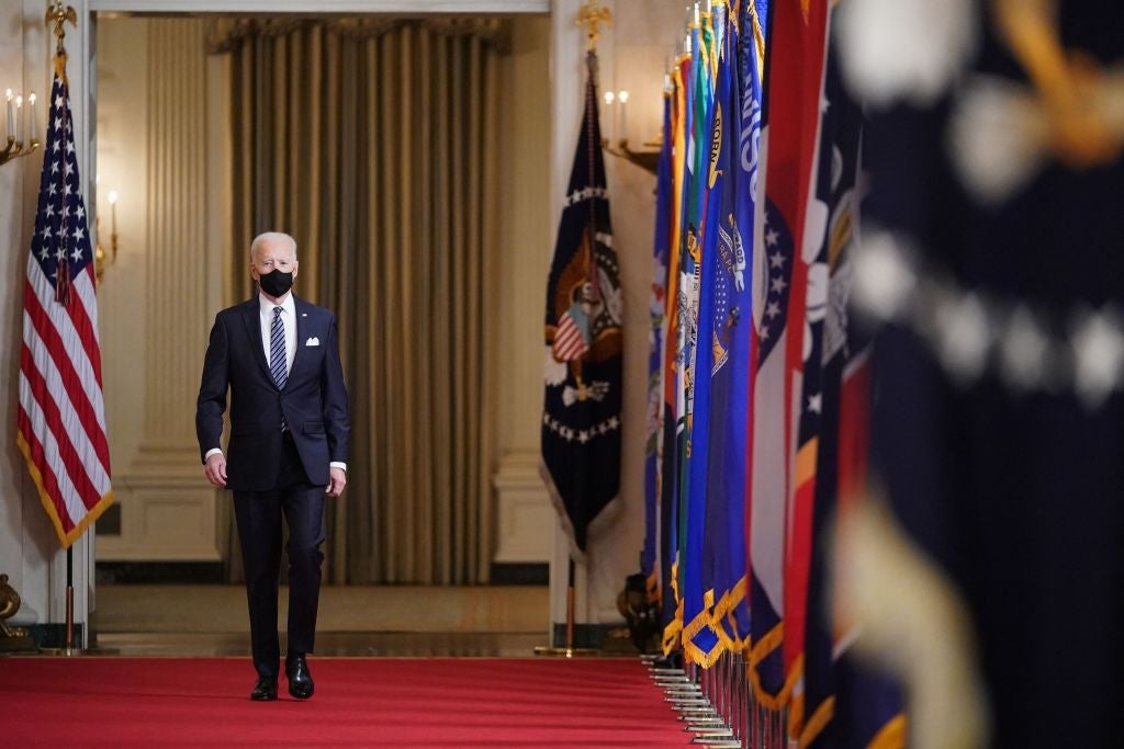 Biden, wearing a suit and a black mask, walks down a red carpet in a hallway lined with flags.