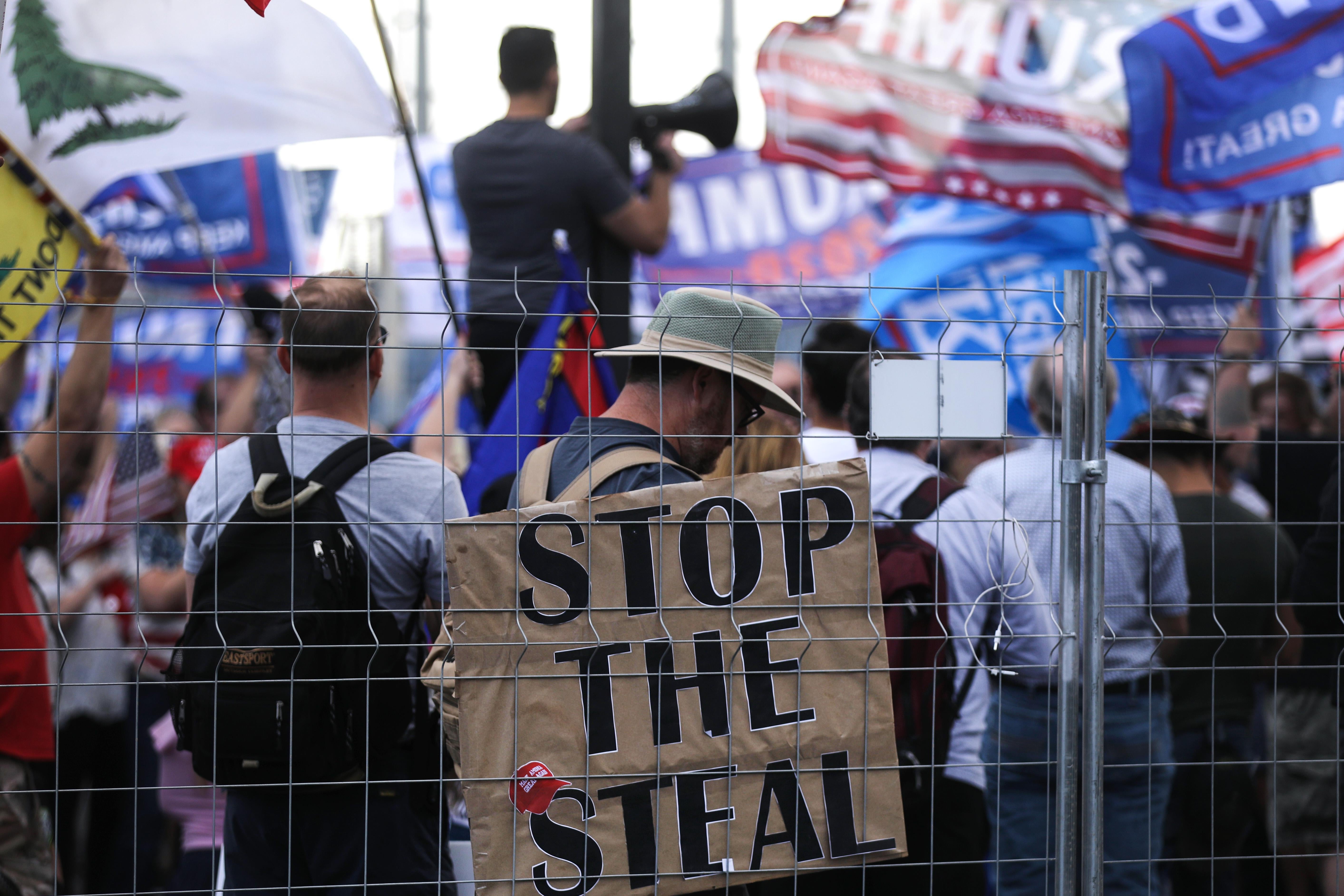 A crowd stands behind a fence with Trump flags and a sign that says "Stop the Steal."