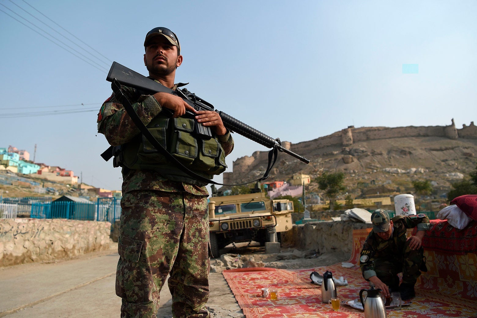 An Afghan soldier stands outside, holding a gun, while another soldier bends down to tend to a tea set on a rug nearby. A military vehicle is in the background.