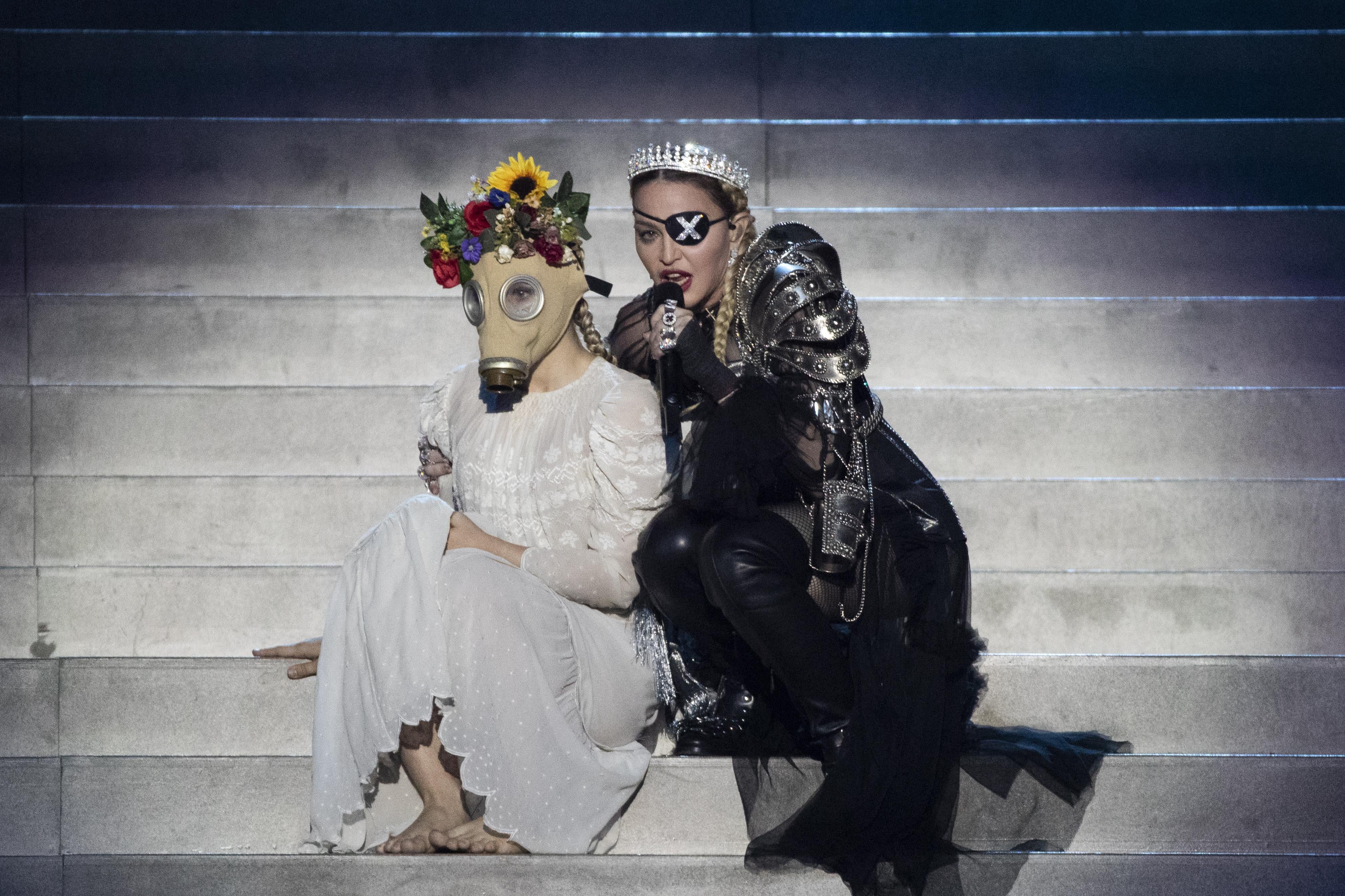 Madonna performs onstage with a dancer wearing a dress, flowers in her hair, and a vintage gas mask.