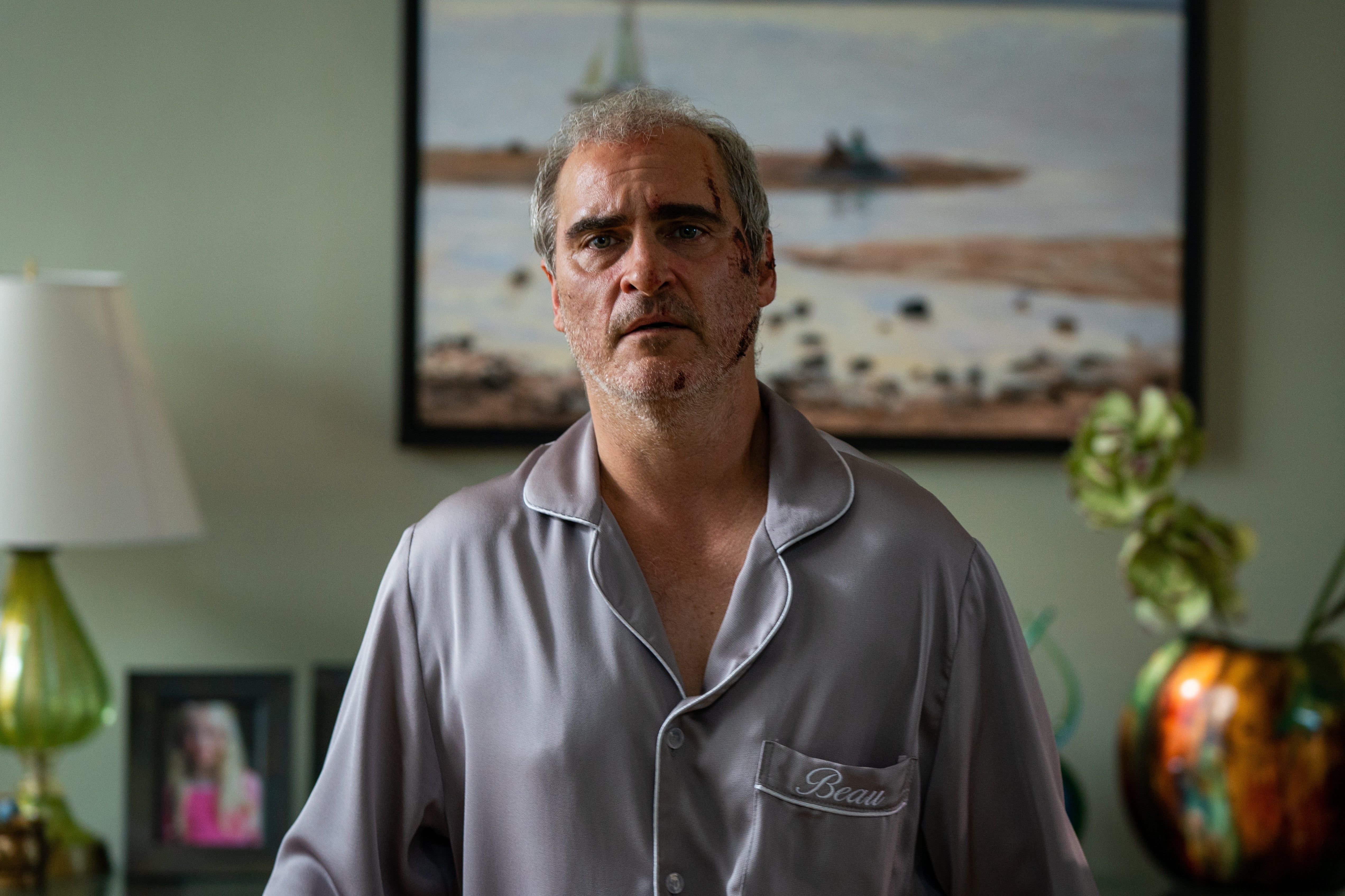A scraped up Joaquin Phoenix stands in silk pajamas labeled "Beau." He looks confused.