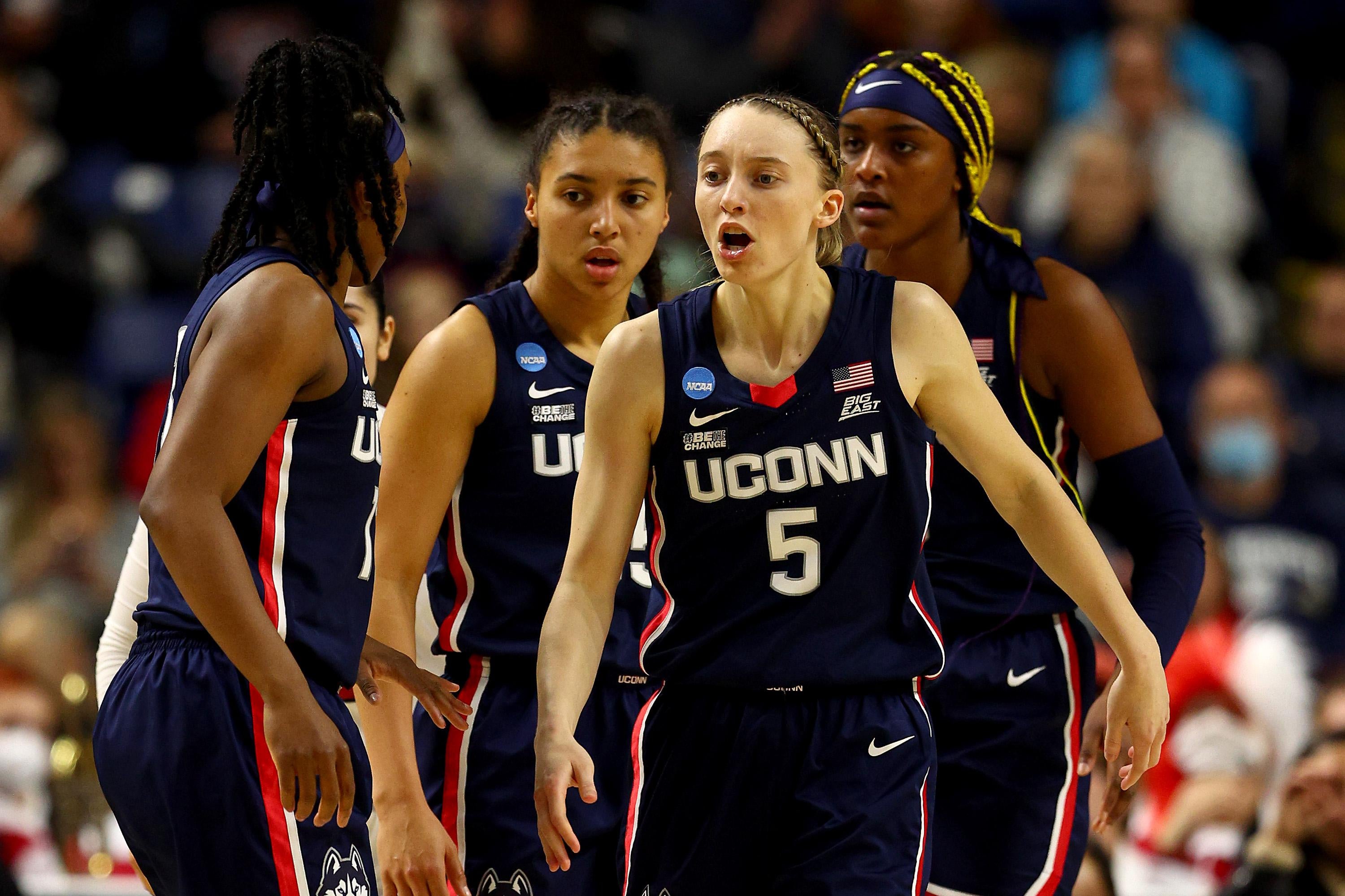 Four UConn women's basketball players standing together on the court wearing navy blue jerseys with a red and white pinstripe.