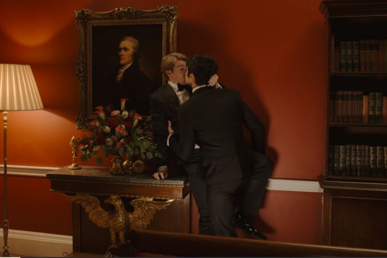 Alex hoists Henry (both in tuxedos) on top of a decorative side table as they make out below a portrait of Alexander Hamilton in the Red Room. 