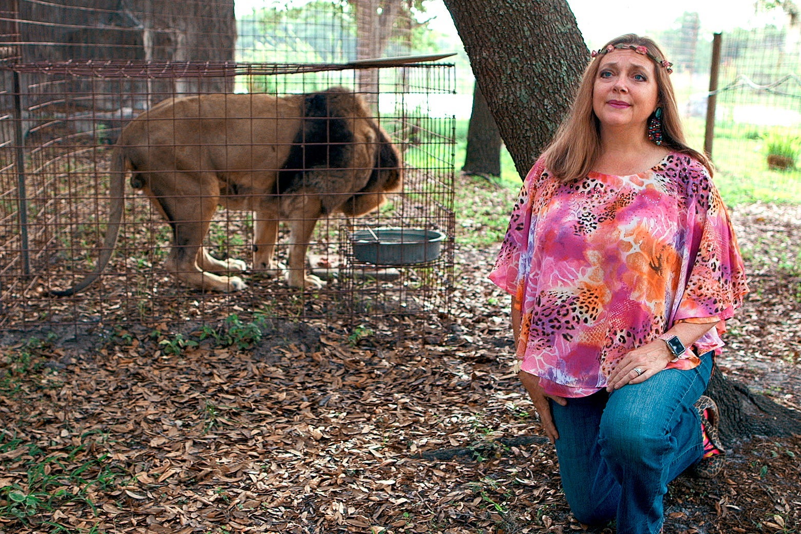 Carole Baskin, wearing a colorful shirt, stands next to a big cat in a wire cage.