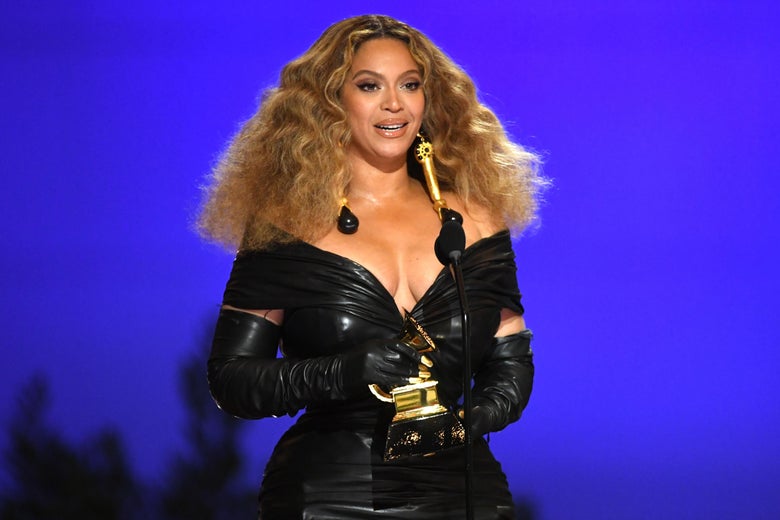Beyoncé, wearing a black dress, stands in front of a blue background holding a Grammy.