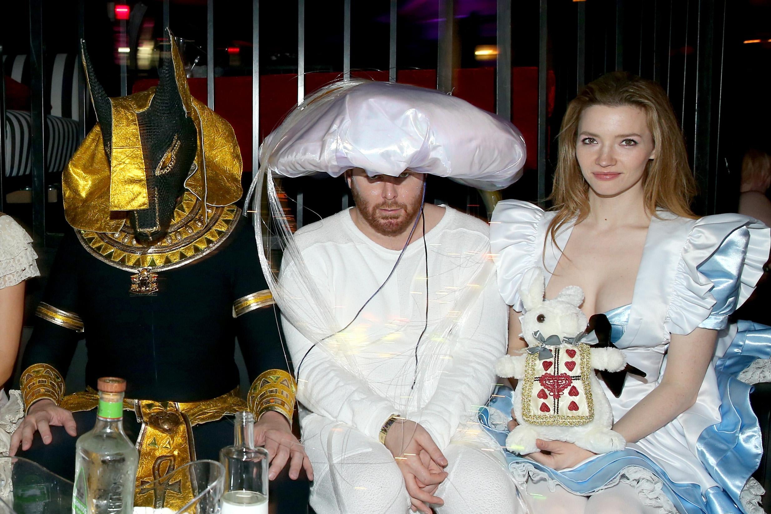 Three people wearing costumes sitting next to each other: one is dressed like an ancient Egyptian deity, one is dressed in all white with a large puffy white hat, and one is dressed like Alice in Wonderland.