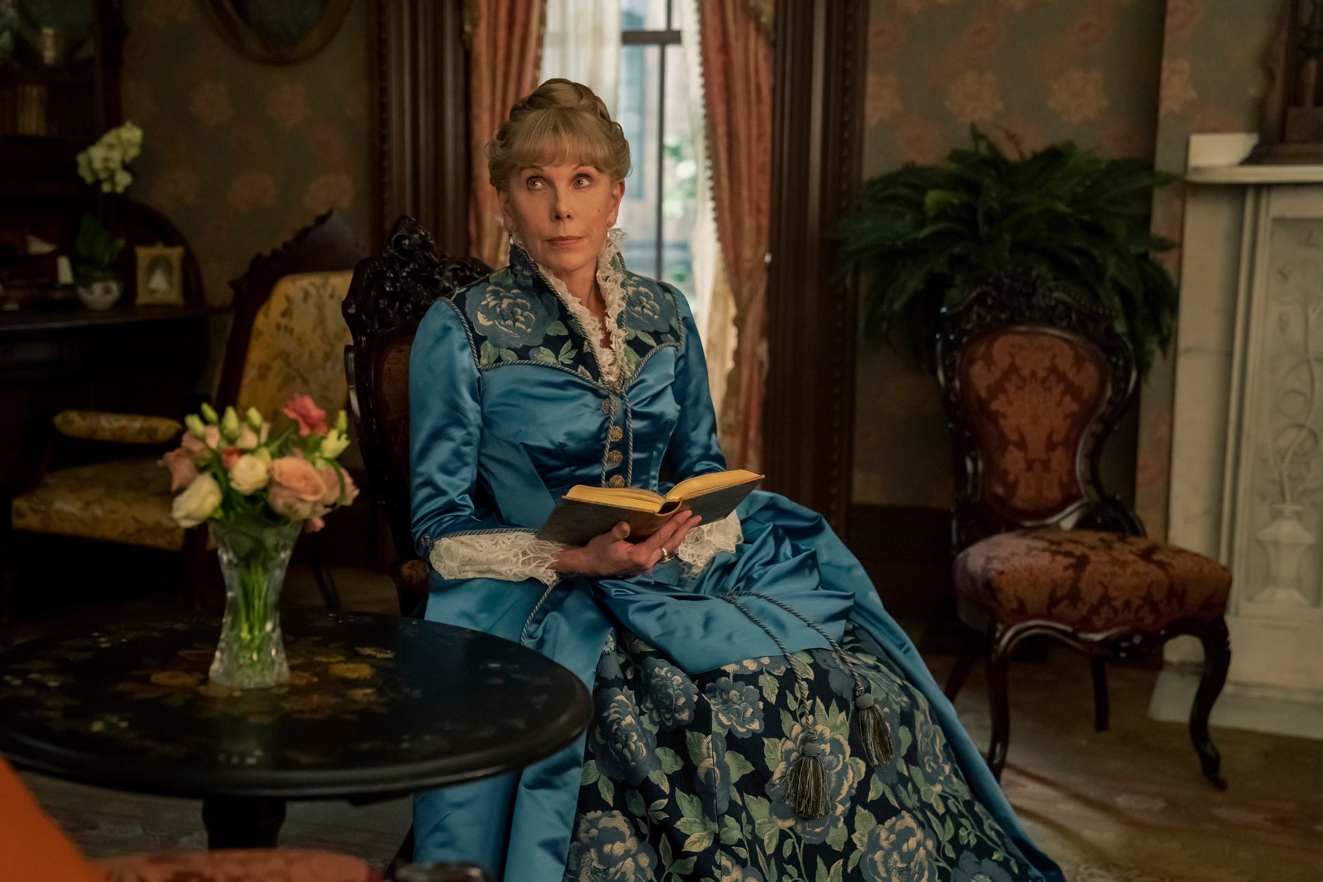 Christine Baranski as Agnes wearing a gorgeous blue dress, seated, with a book in hand.