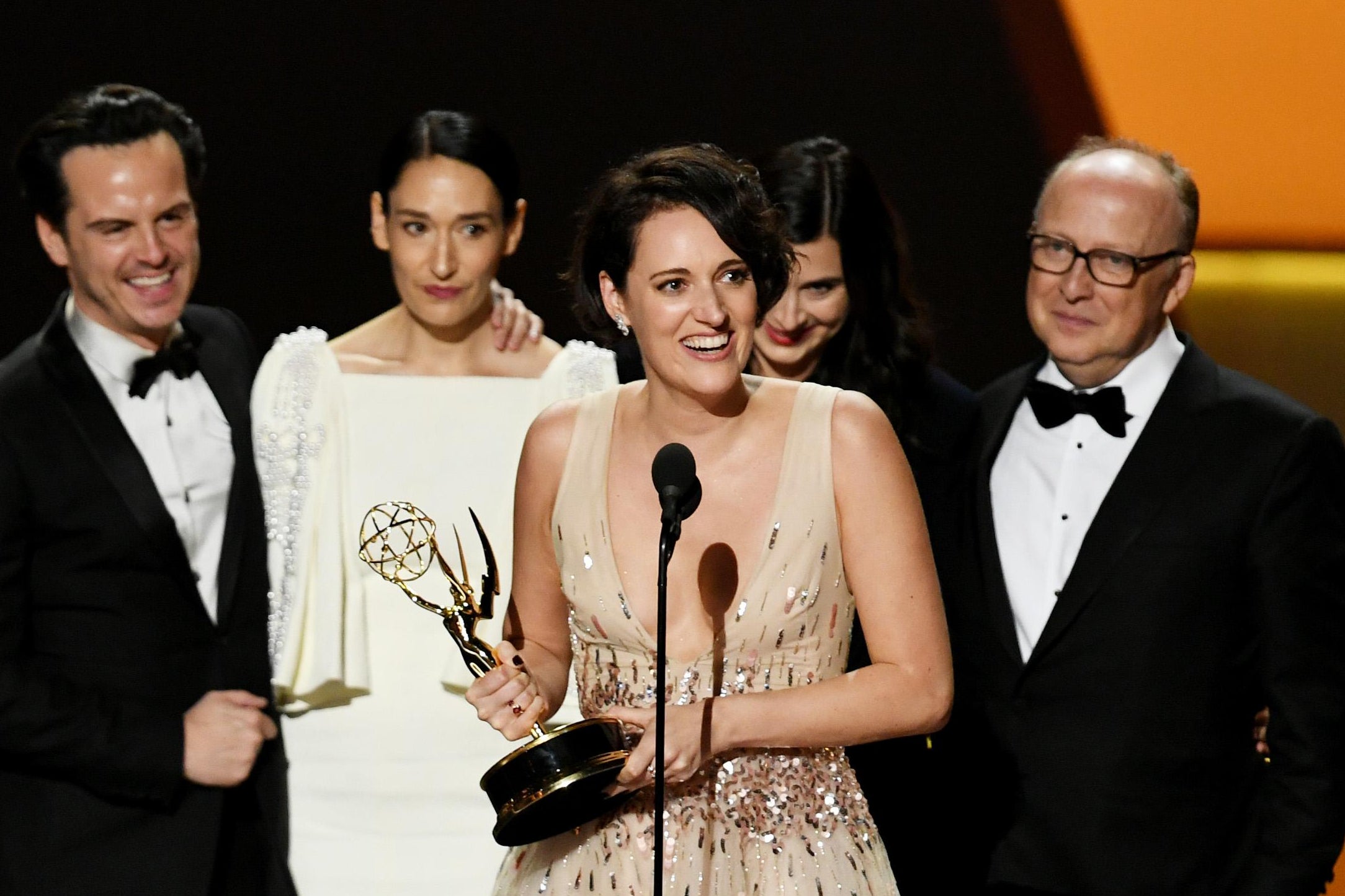 Phoebe Waller-Bridge holds a statuette and speaks into a microphone, surrounded by Fleabag castmates and crew.