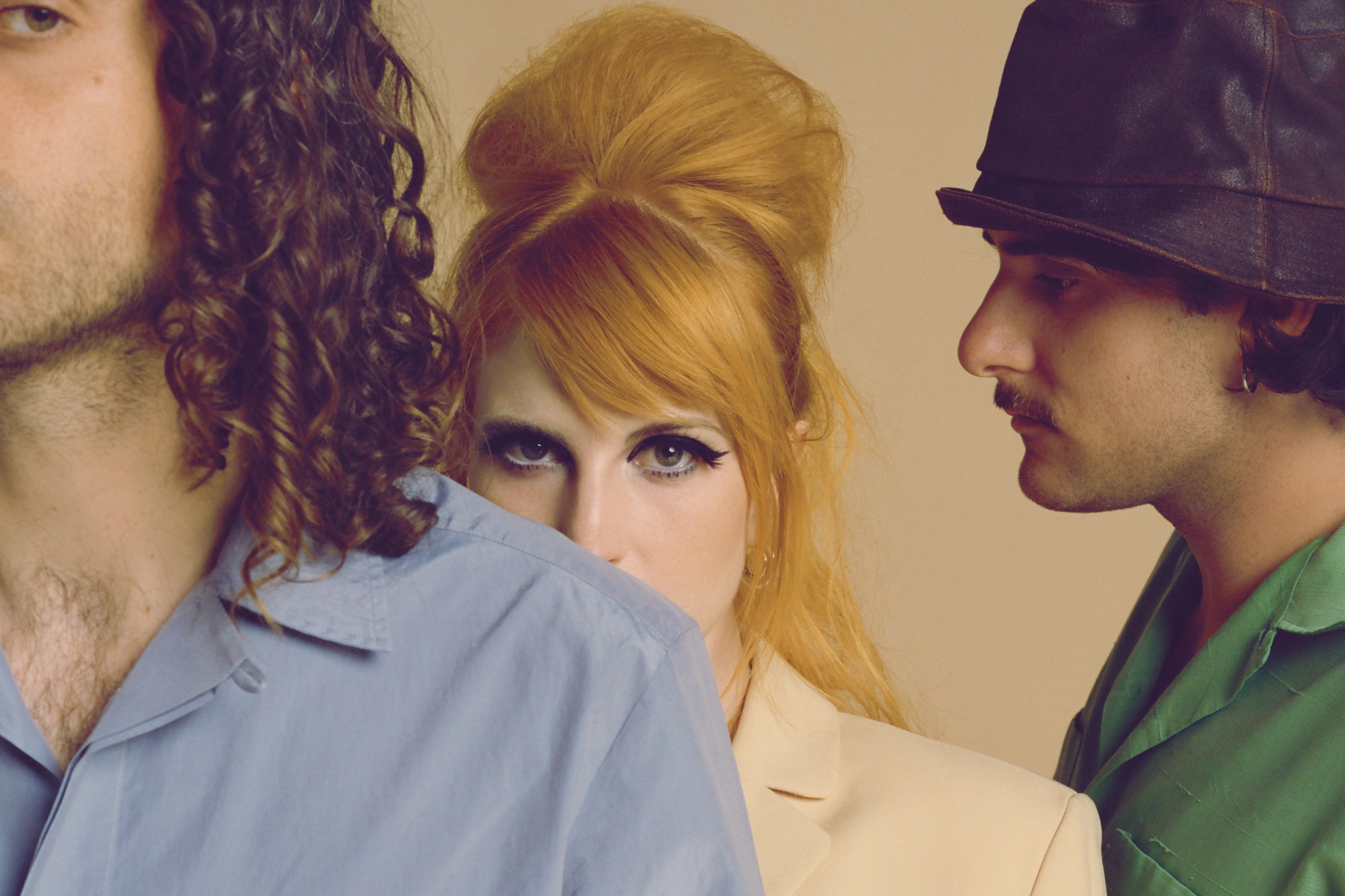 The band Paramore stands in front of a beige colored background. On the left: Taylor York wears a light blue shirt as he faces the camera with half of his face out of frame, center: Hayley Williams wears a peach colored blazer as she stands behind York and stares directly at the camera, the bottom half of her face hidden by York's shoulder, on the right: Zac Farro looks away from the camera, showing his side profile, in a green shirt. 